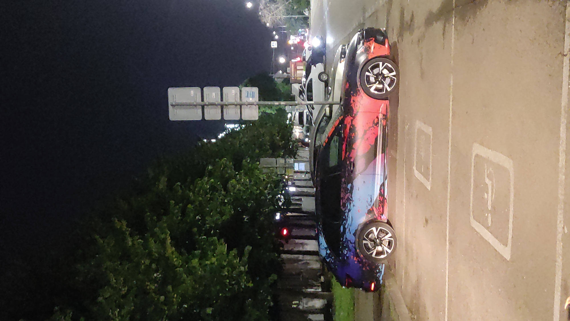 A black Audi A4 Avant with a custom red, blue, and white abstract graphic wrap is parked in a disabled parking spot at night. The car is facing left and is illuminated by streetlights and the headlights of other vehicles. In the background, a traffic light is visible, as are several other cars and trees lining the street. The parking lot is paved and has white lines marking the parking spaces. There are no people visible in the image. The scene appears to be in a city in Krasnodar, Russia, as indicated by the context.