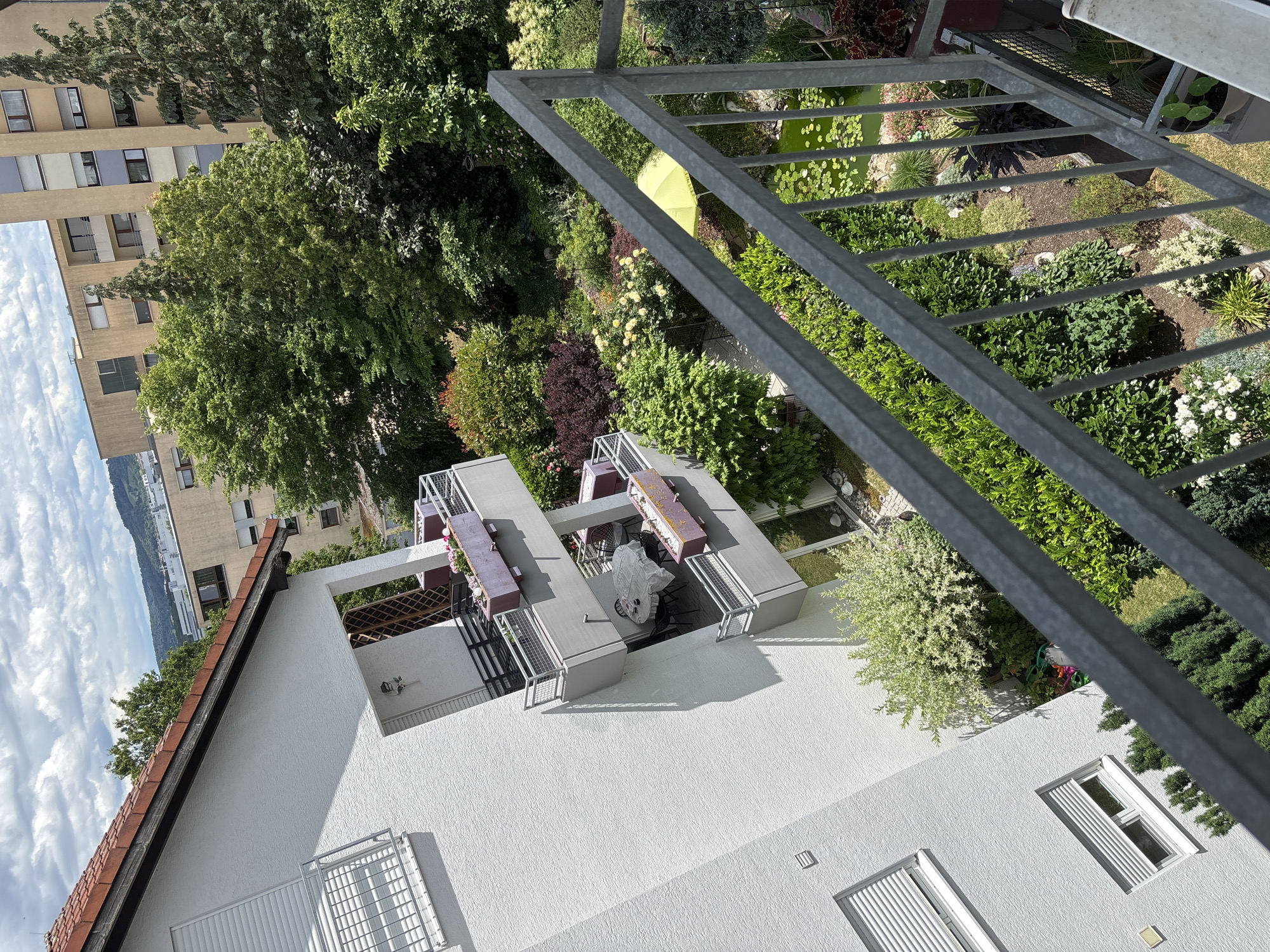 The image captures an elevated daytime view of a residential area in the city of Unknown, Unknown, looking down onto a garden and across to other buildings. In the immediate foreground, a grey metal balcony railing extends diagonally from the lower right to the center. To the left, a multi-story white building with a textured facade features multiple balconies, each equipped with grey metal railings and rectangular planter boxes containing flowers, some purple or pink. One mid-level balcony displays a round table covered by a white tablecloth, surrounded by four dark chairs. Windows with white roller shutters are visible on the building's side, with a terracotta-tiled roof at the top left.
Below and to the right of this building lies a meticulously cultivated garden, showcasing diverse flora including green shrubs, trees, and dark red foliage plants. A small pond with green water and water lilies occupies the lower right section of the garden. A bright yellow or lime green patio umbrella is open, positioned near the pond. In the mid-ground, beyond the garden, several multi-story buildings with beige facades and numerous windows are interspersed with mature green trees. The distant background reveals vegetated hills under a partly cloudy sky. The scene is brightly illuminated, casting clear shadows indicative of daylight. No people or discernible actions are present.