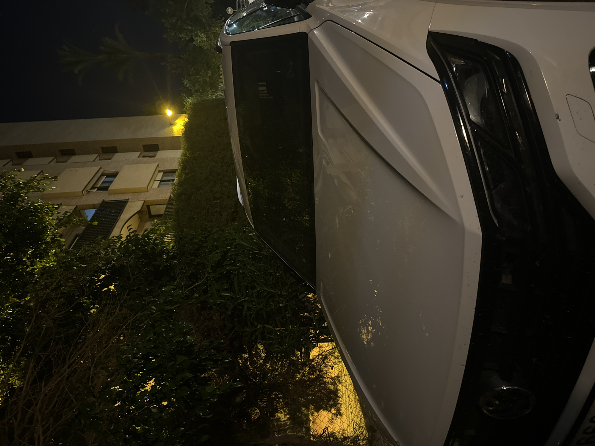 The image shows the front of a white Volkswagen SUV parked at night on a street in Murcia, Spain. The car is white with a black grille and dark tinted windows. The headlights are visible and appear to be on, casting a slight glow. In the background, a multistory building with balconies and lit windows is visible, along with trees and foliage. A street lamp with a yellow light illuminates a portion of the scene. The overall impression is of a residential street at night, with the car parked in front of a building.