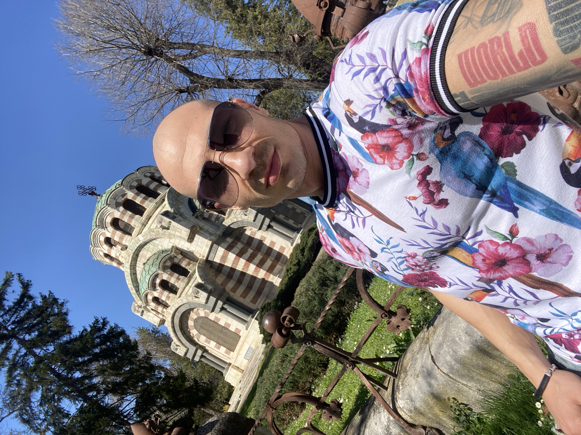 A selfie shot outdoors in Pleven, Bulgaria, features a bald man wearing sunglasses and a floral-patterned shirt. The man has tattoos visible on his arm, including one that says "WORLD." In the background, a section of the Church of St. George the Victorious (Mausoleum) can be seen, recognizable by its architectural design. The scene also includes greenery, trees, and an ornate, rustic metal fence. The sky is clear and blue, suggesting a sunny day.