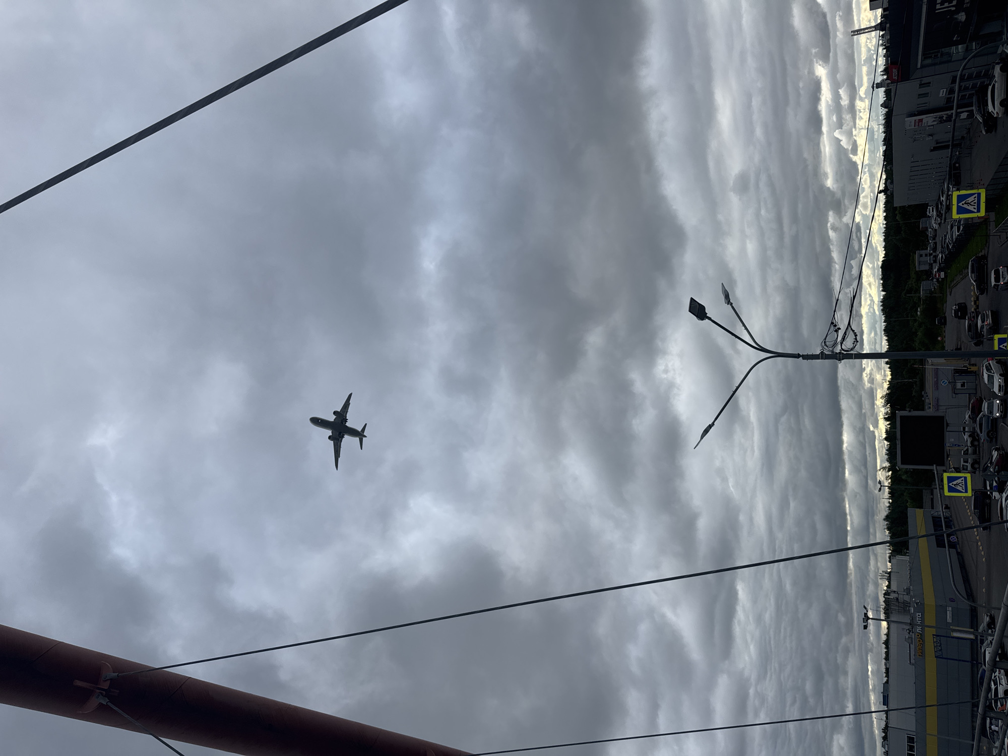The image shows a cloudy sky with an airplane in silhouette, seen from below. The airplane is in the upper left quadrant of the frame, and its wings are extended. A dark, curved object, likely a structure or building element, occupies the bottom left corner. Several dark wires crisscross the sky. In the background, to the right, there is a cityscape with buildings and a parking lot filled with cars. A street light with three lamps is visible in the middle right of the frame. Two yellow pedestrian crossing signs with blue diamond shapes are also present. The sky is overcast, with dramatic, textured clouds. The overall impression is of an overcast day, possibly late afternoon or early evening, with an urban setting. There is some text visible on a building in the background, appearing to be in Cyrillic script.