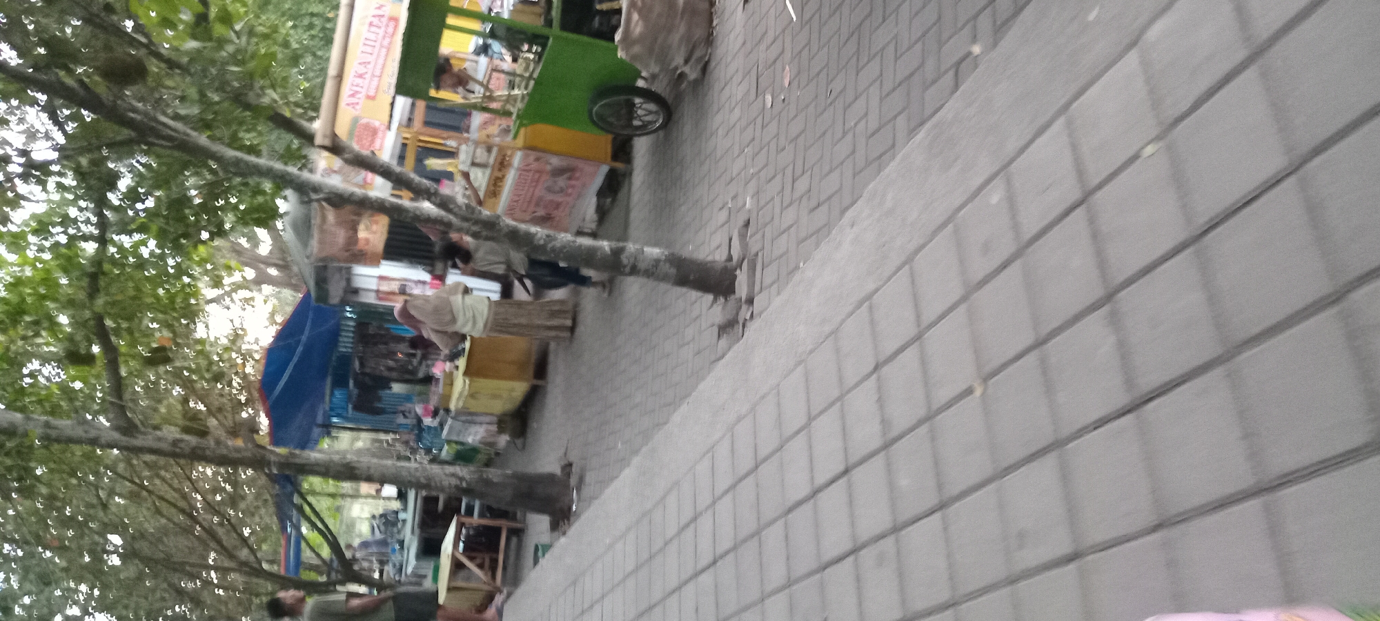 The image depicts an outdoor scene at a market or street food area, likely in Mataram, Indonesia. In the foreground, a paved walkway with rectangular gray tiles stretches into the distance. To the right of the walkway, a green food cart with a bicycle wheel is visible. Behind the cart, a sign reads "ANEKA LILITAN". Further back, several food stalls with blue and yellow awnings are set up. People are seen browsing or working at these stalls. One person, a woman wearing a hijab and a long skirt, appears to be at one of the stalls. Another person, a young girl, stands near the green food cart. A man wearing a green shirt and shorts is visible on the left side of the frame, standing near a wooden table. Trees with green foliage are interspersed throughout the scene, providing shade. The overall impression is a bustling market environment during daylight hours.