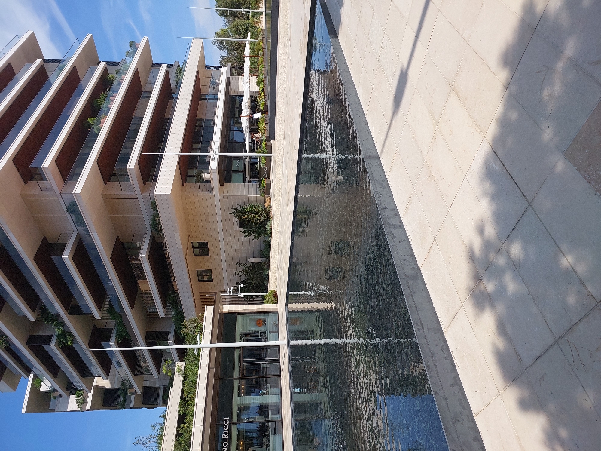 The image shows a modern, multi-story building with a light-colored facade and numerous balconies with glass railings. The building is situated in an urban environment, likely Monaco, given the architecture and the presence of a public plaza with a reflecting pool in the foreground. The pool is dark and still, reflecting the building and the sky, which is clear and blue. The plaza is paved with light-colored tiles, and shadows from unseen trees are cast on the ground. On the ground level of the building, there appear to be shops or restaurants with large glass windows. One shop has the name "NO RICCI" visible. Several outdoor umbrellas are set up in front of what looks like a cafe or restaurant area. There are no people clearly visible in the image, but the presence of umbrellas and the storefronts suggest a public area that might be frequented by people. The overall impression is one of contemporary architecture and urban design, creating a serene and upscale atmosphere.