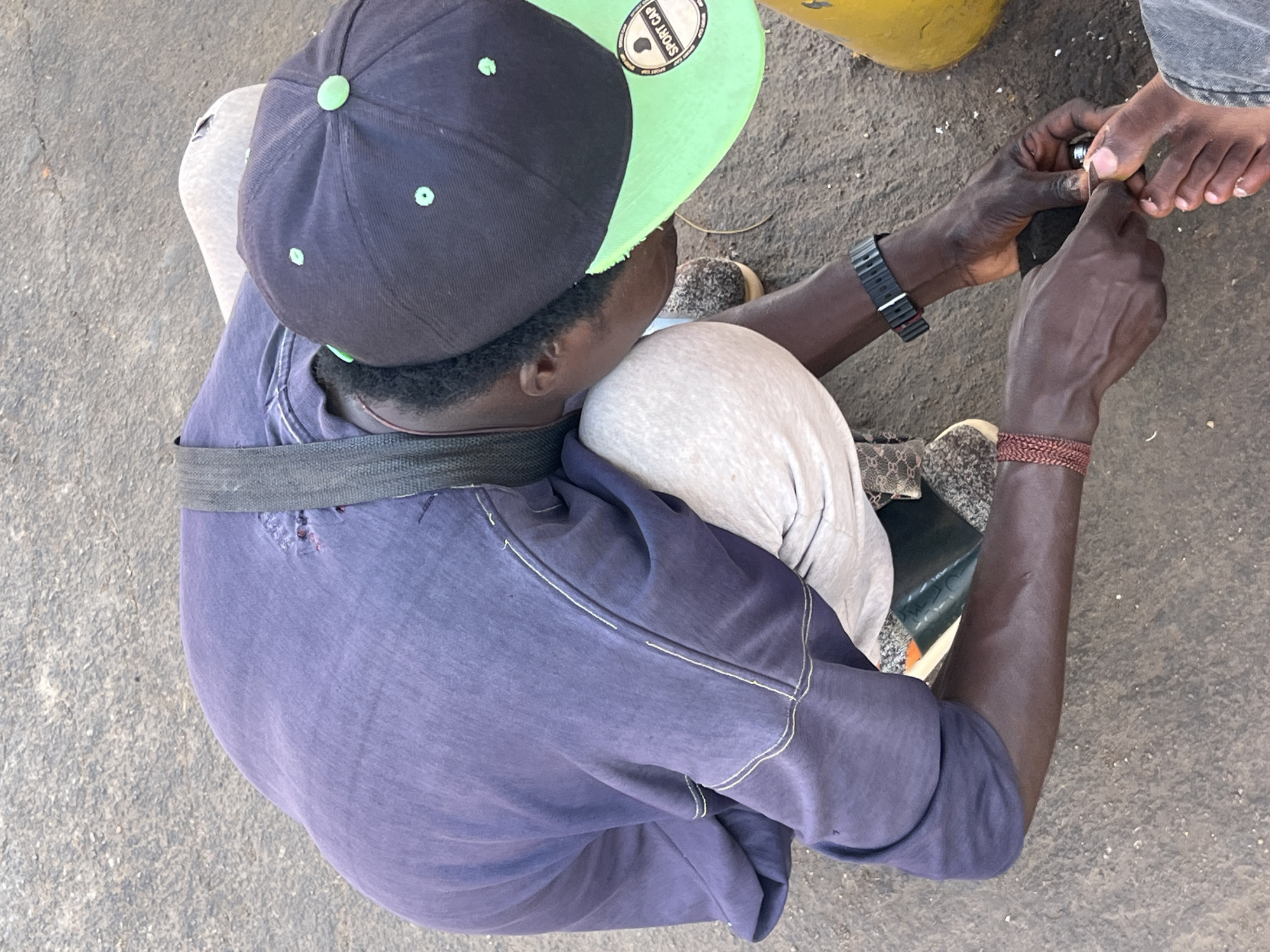 An overhead shot captures an outdoor scene in Jos, Nigeria, showing a person providing nail care. The individual, who has dark skin, is seated or kneeling on a grey, possibly concrete or dirt, ground surface. They are wearing a black baseball cap with a prominent neon green brim, on which the text "SPORT CAP" and a small logo are visible. Their attire includes a faded purple short-sleeved shirt with white stitching details and a visible tear on the back, along with light grey pants.
The person's hands are focused on trimming the toenails of another individual, whose bare feet are visible in the upper right. They hold a silver nail clipper or similar tool. Accessories worn by the person providing the service include a black watch with a dark band on their left wrist and a woven brown bracelet on their right wrist. A dark grey or green strap is also visible across their lower back/waist area. Part of a yellow object can be seen in the upper right background. The scene is brightly lit, indicating it is daytime.