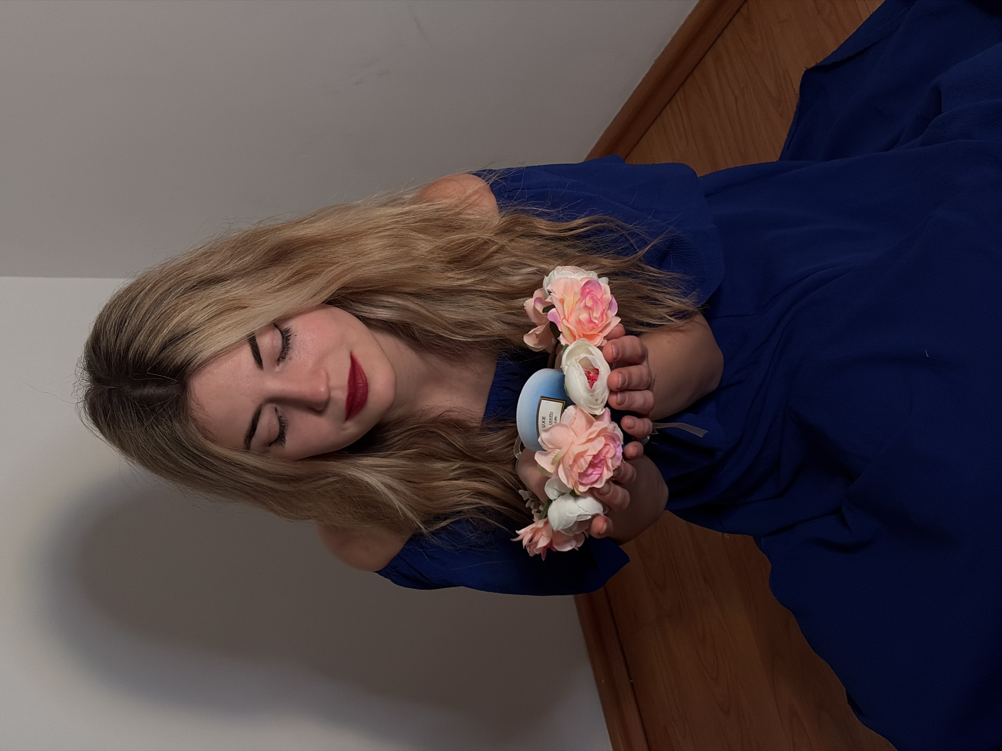 A young woman with long, wavy blonde hair is depicted in a close-up, portrait-style shot. She is positioned as if reclining or lying down, with her head slightly tilted. Her eyes are closed, and she has a calm, serene expression on her face, accented by dark red lipstick. Her eyebrows are dark and well-defined.
She is wearing a dark blue, off-the-shoulder dress, the fabric of which appears soft and flowing, with some gathering around the neckline. Her bare shoulders are visible.
In her hands, which are held close to her upper chest/shoulder area, she is holding a small collection of artificial flowers. These flowers are in shades of light pink, peach, and white, resembling peonies or roses. Nestled amongst the flowers, she also holds a small, light blue, cylindrical object. Visible text on this object reads "LUXE" at the top, and "seaside" below it, suggesting it might be a small candle or scented item.
The background consists of a plain, light-colored wall on the left side of the frame, and a wooden floor with distinct grain and color variations on the right side. Soft shadows are present on the wall, indicating indoor lighting. The overall mood of the image is peaceful and contemplative.