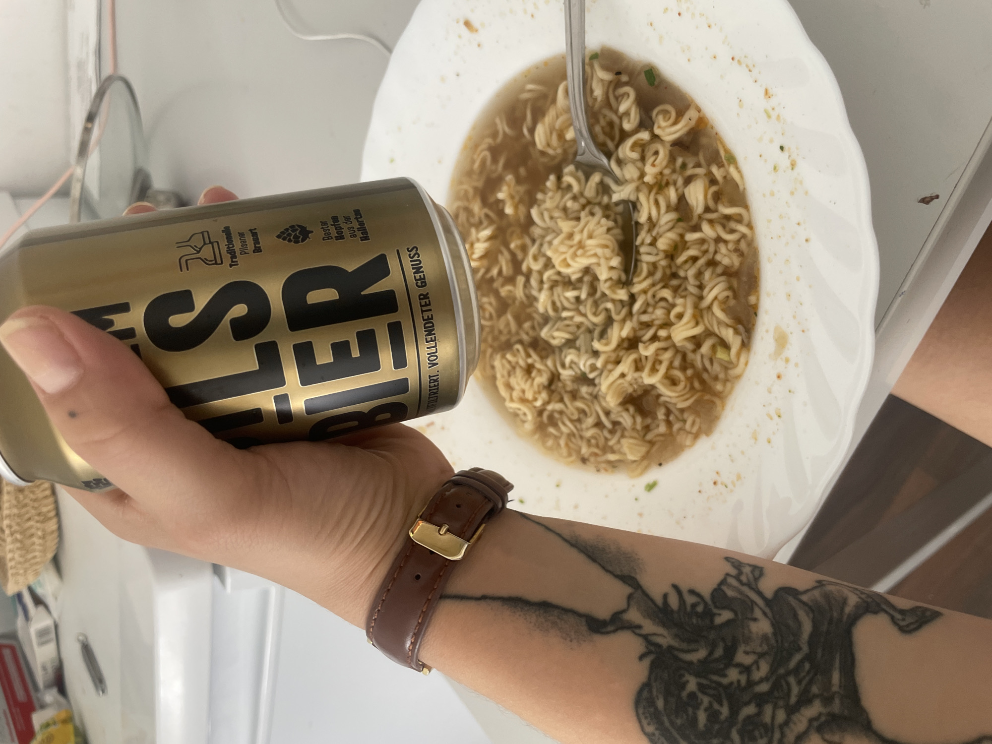 The image shows a person holding a can of beer over a bowl of ramen noodles. The person has a tattoo on their arm and is wearing a brown leather watch. The scene appears to be indoors, possibly in a home kitchen. There is no indication of the location being in the city of Solingen, Germany.