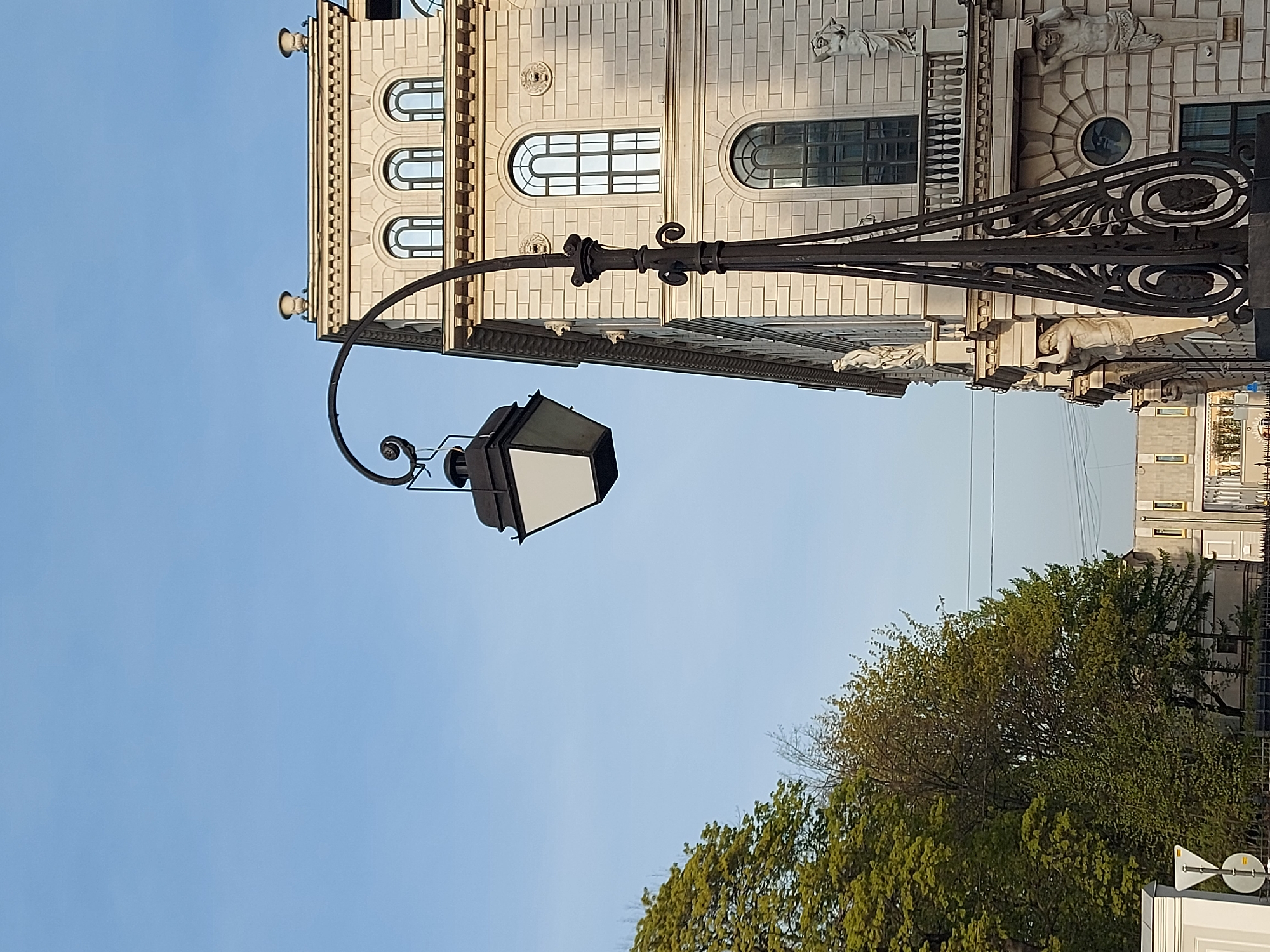 The image shows a street lamp in Saint Petersburg, Russia. The lamp has a black metal frame with a white, box-shaped light fixture. It extends out from a building with ornate architectural details, including windows, statues, and decorative molding. The building is made of light-colored stone or brick. Below the lamp and building, there are green trees and a clear blue sky.