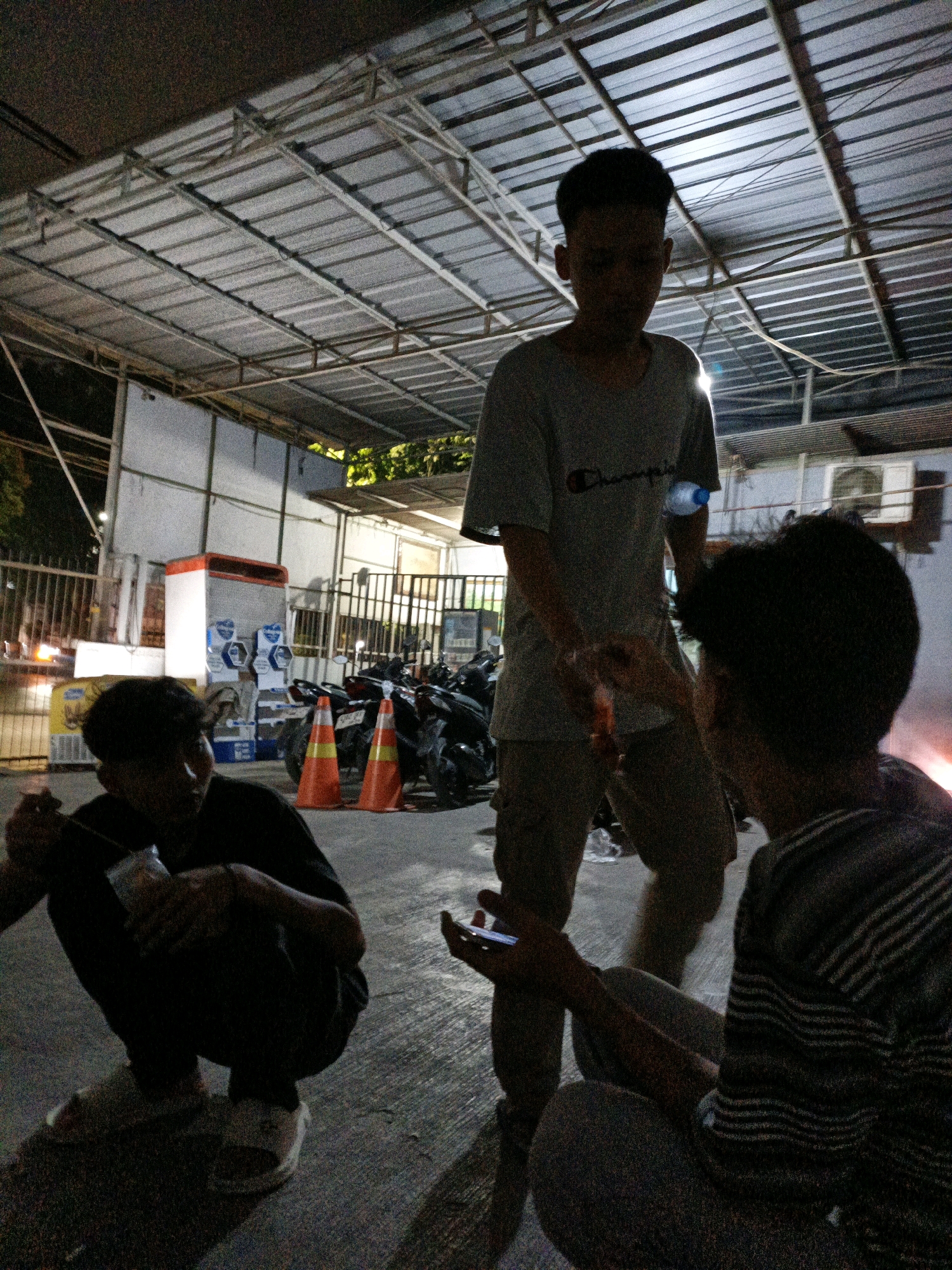 The image depicts an outdoor scene at night, possibly a street food stall or a casual gathering area in North Jakarta, Indonesia. Three young men are visible, silhouetted against the dim lighting. One man stands in the center, wearing a t-shirt and shorts, holding a water bottle. To his left, another man is crouched down, holding a food item. To the right, a third man is seated, looking at his phone. In the background, there are parked motorcycles, traffic cones, and what appears to be a vending machine or kiosk. The overall atmosphere suggests a relaxed, informal social gathering.