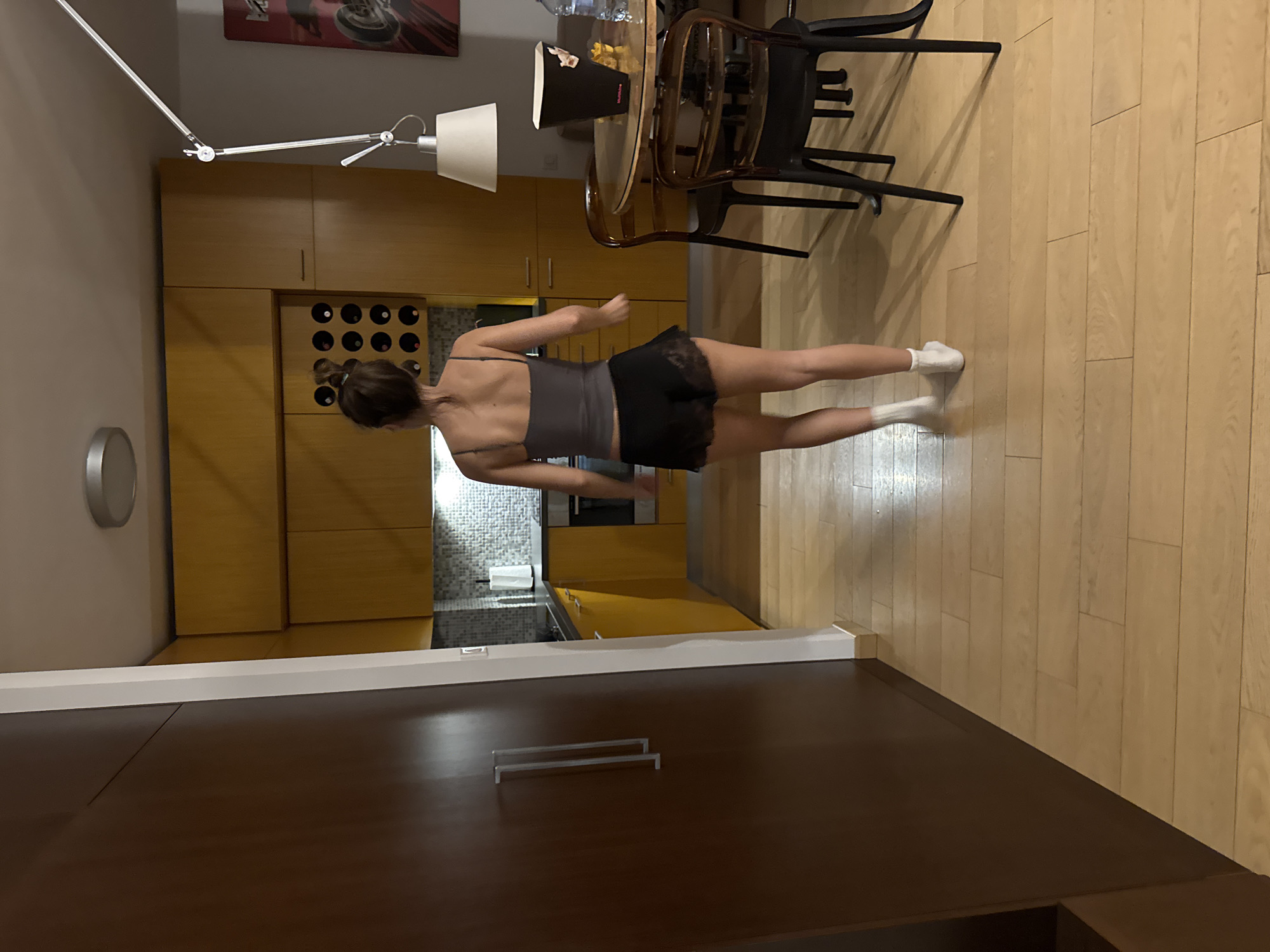 A photograph taken indoors shows a woman from behind, standing on a wooden floor in a room with modern decor. She is wearing a gray tank top and dark shorts with white socks. In the background, there are light-colored wooden cabinets and a dining area with a table and chairs. The scene is lit by a modern overhead lamp. The overall impression is that of a contemporary living space, possibly located in Warsaw, Poland, although there are no features within the image that specifically establish this.