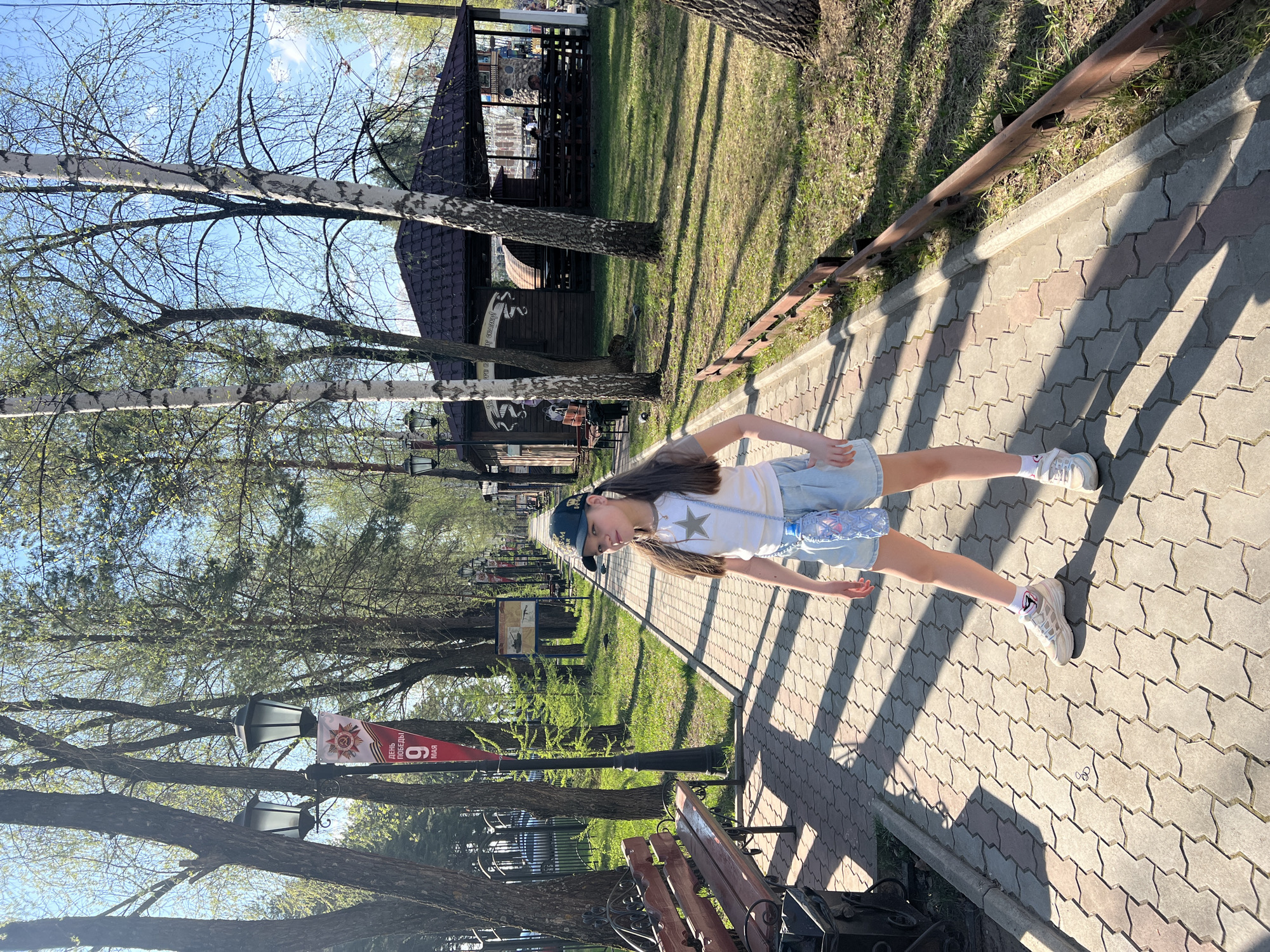 A young girl stands on a cobblestone pathway in Chaikovsky, Russia, on a sunny day. She wears a white T-shirt with a star, denim shorts, sneakers, and a baseball cap, and she has a water bottle hanging from her shoulder. The pathway is bordered by a rusty rail, grass, and trees. Behind the girl, there are park benches, lamp posts, and a wooden building. A banner with "9 May" written in Russian hangs from a lamppost.