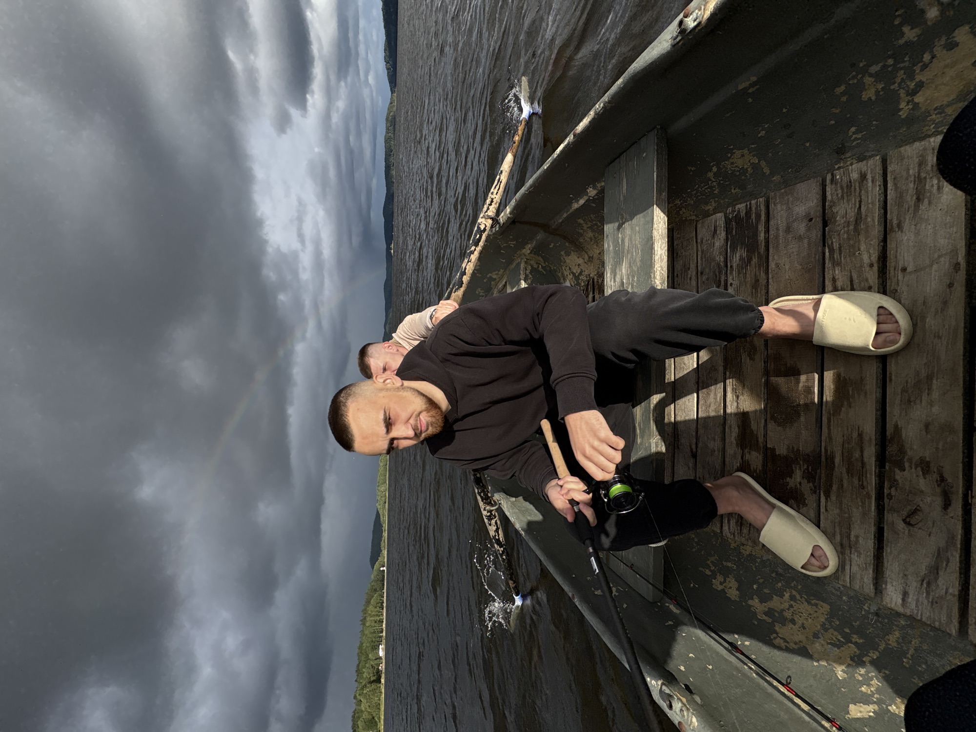 Two individuals are in a rowboat on a body of water. The person in the foreground, seated and facing slightly right, is wearing a black hoodie and dark pants, with light-colored slides on their feet. They are holding a fishing rod. Behind them, another individual is rowing the boat, also facing forward. The boat is made of metal and has wooden seating. The water appears choppy. The background features a treed shoreline and hills under a dramatic, cloudy sky with a visible rainbow. The location is identified as Rautalakhti, Russia.