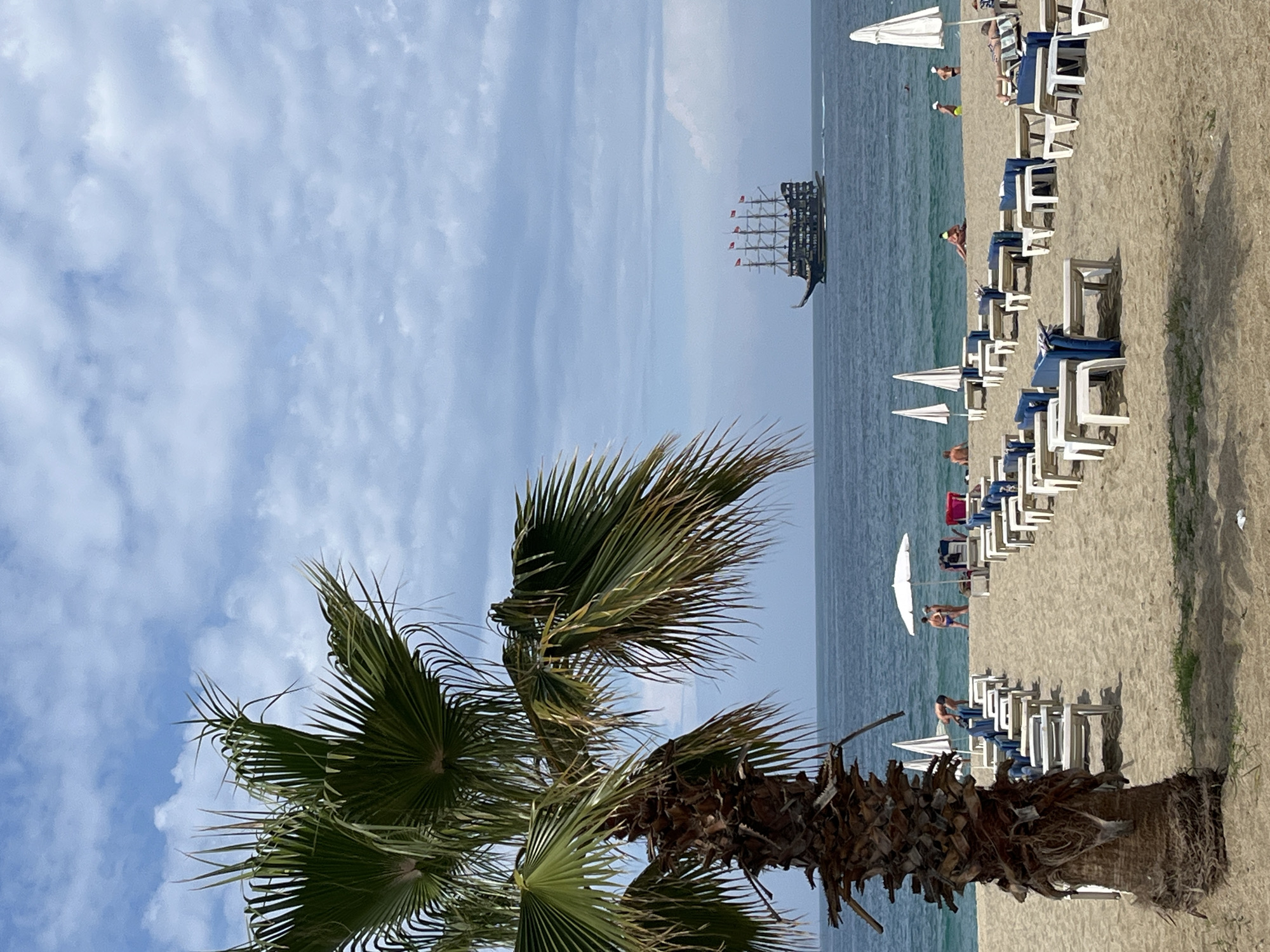 The image shows a beach scene in Alanya, Turkey. A palm tree is prominent in the foreground. Beyond the tree, the view opens to the sea with a large pirate-themed sailing ship visible on the water. The beach is populated with rows of white and blue sun loungers and white umbrellas. People are seen lounging and walking along the beach. The sky is blue with scattered clouds. The overall atmosphere appears calm and sunny.