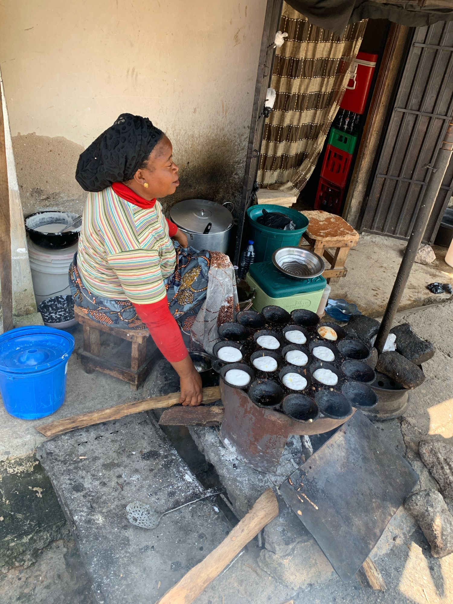 In the city of Jos, Nigeria, a woman is pictured in an outdoor setting, likely a market stall or street-side kitchen. She is wearing a black headscarf and a colorful striped long-sleeved shirt with red cuffs, layered over a red turtleneck. Her patterned skirt is visible as she sits on a wooden stool, facing right. In front of her, a rustic cooking setup is in progress. A cast-iron mold with multiple round indentations is placed over a fire, with several small, round white pastries cooking in it. A long wooden stick is positioned to help manage the cooking process. To the left, a large blue plastic bucket and other containers, including a pot and what appears to be a cooler, are situated. In the background, a striped curtain hangs, and stacks of crates containing beverages are visible. The ground is rough and unpaved, consistent with an outdoor, informal commercial area.
