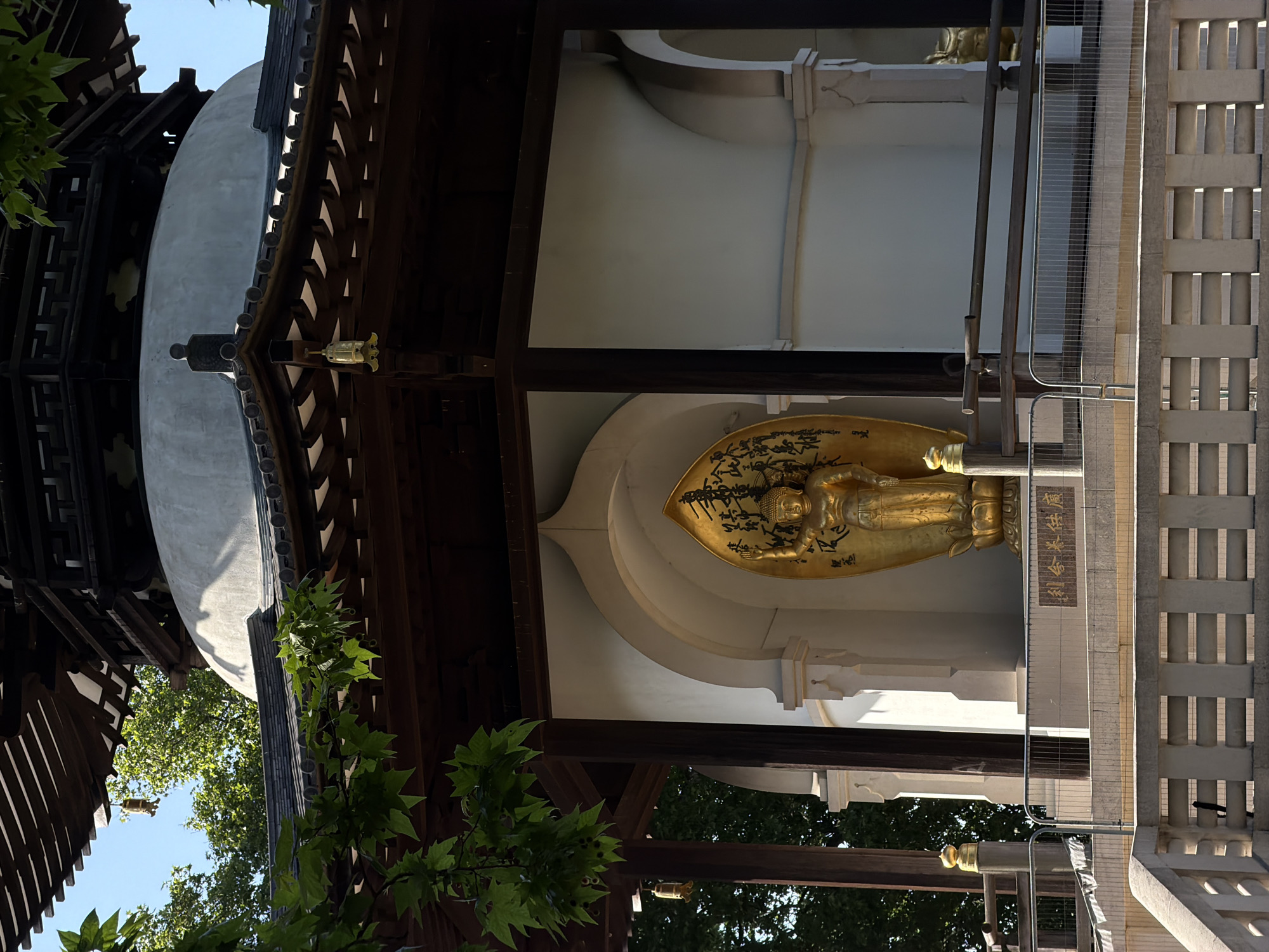 The image depicts a golden reclining Buddha statue within a niche in a traditional Asian-style building. The statue is adorned with what appears to be calligraphy or decorative elements on its surface. The building features dark wood beams and a tiered roof structure. To the side, a gray, rounded structure resembling a dome can be seen. Vegetation is visible in the foreground. A stone pillar-like structure runs adjacent to the building. Based on visual cues such as the building's architectural style, this is likely in a garden or park setting in the city of London, United Kingdom.