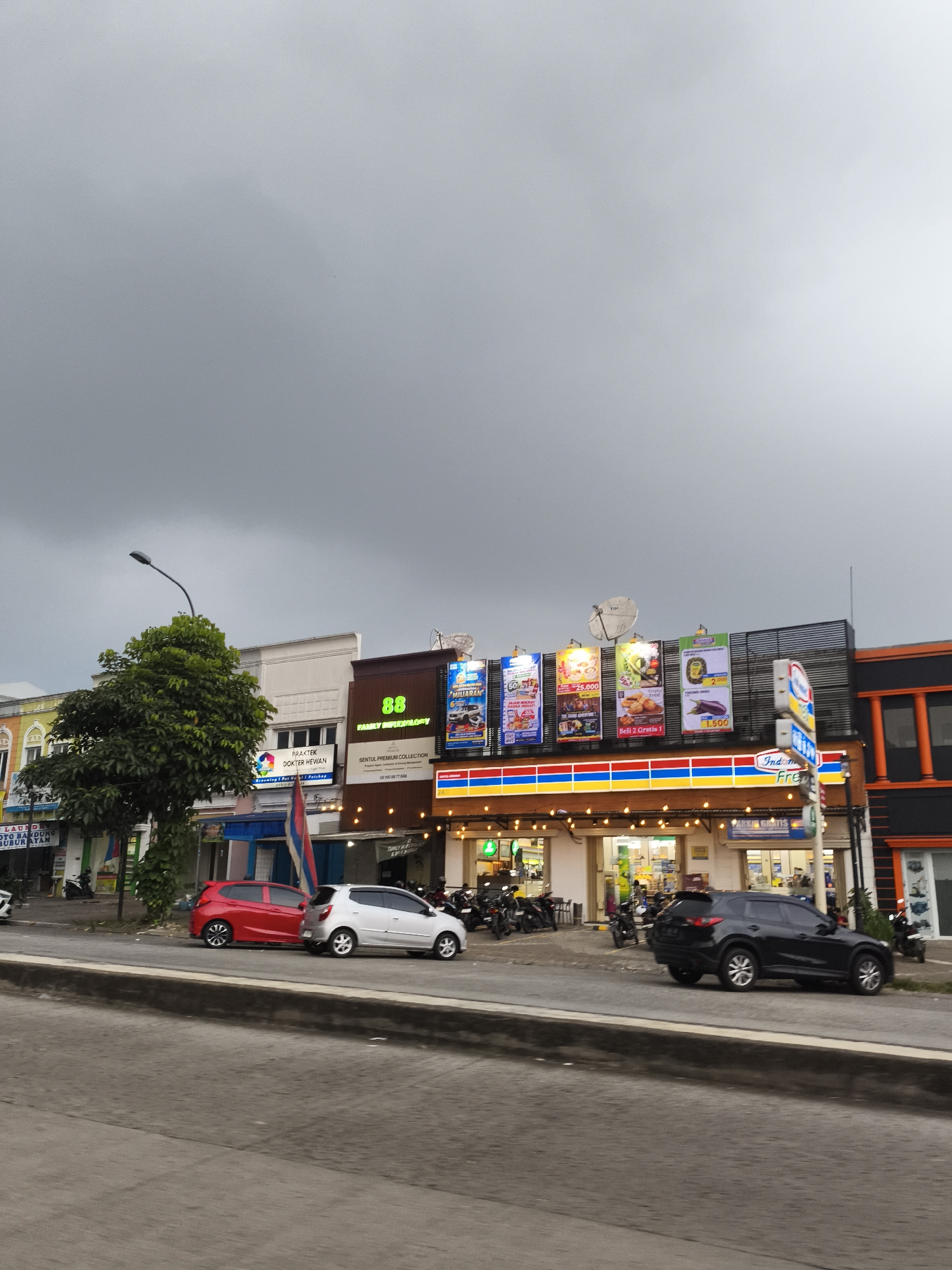 This media file depicts a bustling commercial street in Babakan Madang, Indonesia, under an overcast, cloudy sky. The foreground shows a wide road separated by a low concrete barrier from the sidewalk and a row of commercial buildings.
On the right, a brightly lit Indomaret convenience store is prominent, featuring its characteristic blue, red, and yellow striped facade. Above the store, multiple colorful banners advertise promotions, with visible text including "25.000," "1.500," and "Beli 2 Gratis 1" (Buy 2 Get 1 Free). A partially visible sign indicates "PARKIR GRATIS" (Free Parking) at the Indomaret. Two satellite dishes are mounted on the roofline above the store.
To the left of the Indomaret, buildings house other businesses. One displays a large "88" and "FAMILY INIFLEXOLORY" sign, with another reading "SENTUL PREMIUM COLLECTION." Adjacent to this is a "PRAKTEK DOKTER HEWAN" (Veterinary Practice). Further down the street, signs for "LAUNDI," "TO BANDUNG," and "BUBUR AYAM" (Chicken Porridge) are visible.
Several vehicles are parked along the sidewalk: a red hatchback, a white hatchback, and a black SUV. Numerous motorcycles and scooters are also parked, suggesting active businesses. A street lamp and a large green tree stand on the left side of the street. The scene captures an everyday commercial environment during daytime, characterized by a mix of local businesses and a major convenience store.