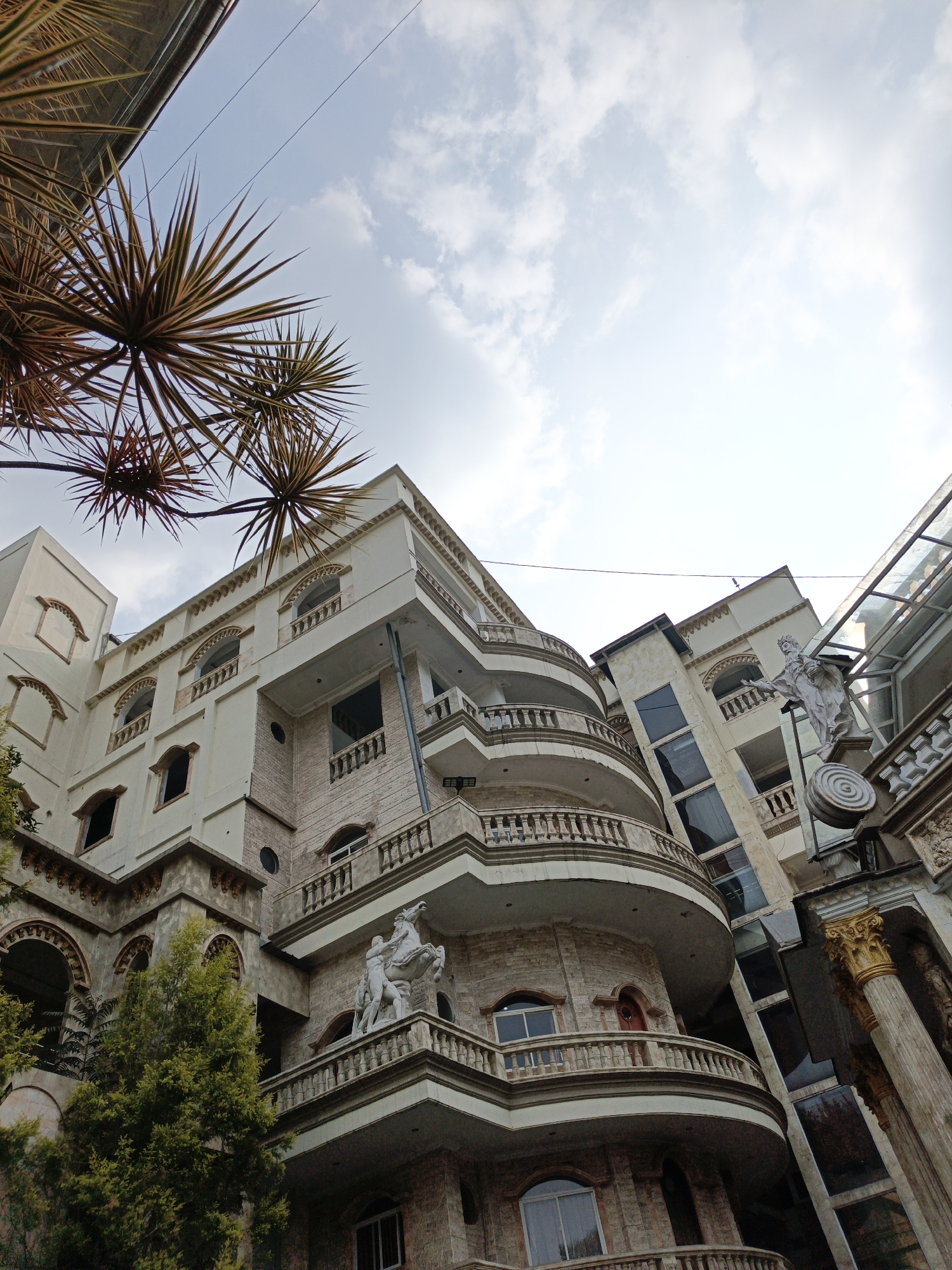 This is a low-angle, full-shot photograph of a grand, multi-story building with an ornate architectural style. The building is constructed with a light-colored facade and accents of stone and gold-colored columns. It features multiple curved balconies with decorative railings, arched windows, and various sculptural elements. Notably, there is a white statue of two figures, one on horseback, displayed on a balcony.
In the foreground, the spiky fronds of a plant are visible against a light blue sky with scattered white clouds. The building itself appears to have a mix of modern and classical design elements, including large windows and what seems to be a glass-enclosed elevator shaft. The overall impression is one of opulence and grandeur.
The location is identified as Cibogo, Indonesia. The scene depicts a static, architectural subject, with no people or animals actively present. The environment is clearly urban, with the building being the central focus. The lighting suggests it is daytime, possibly late afternoon given the soft light.