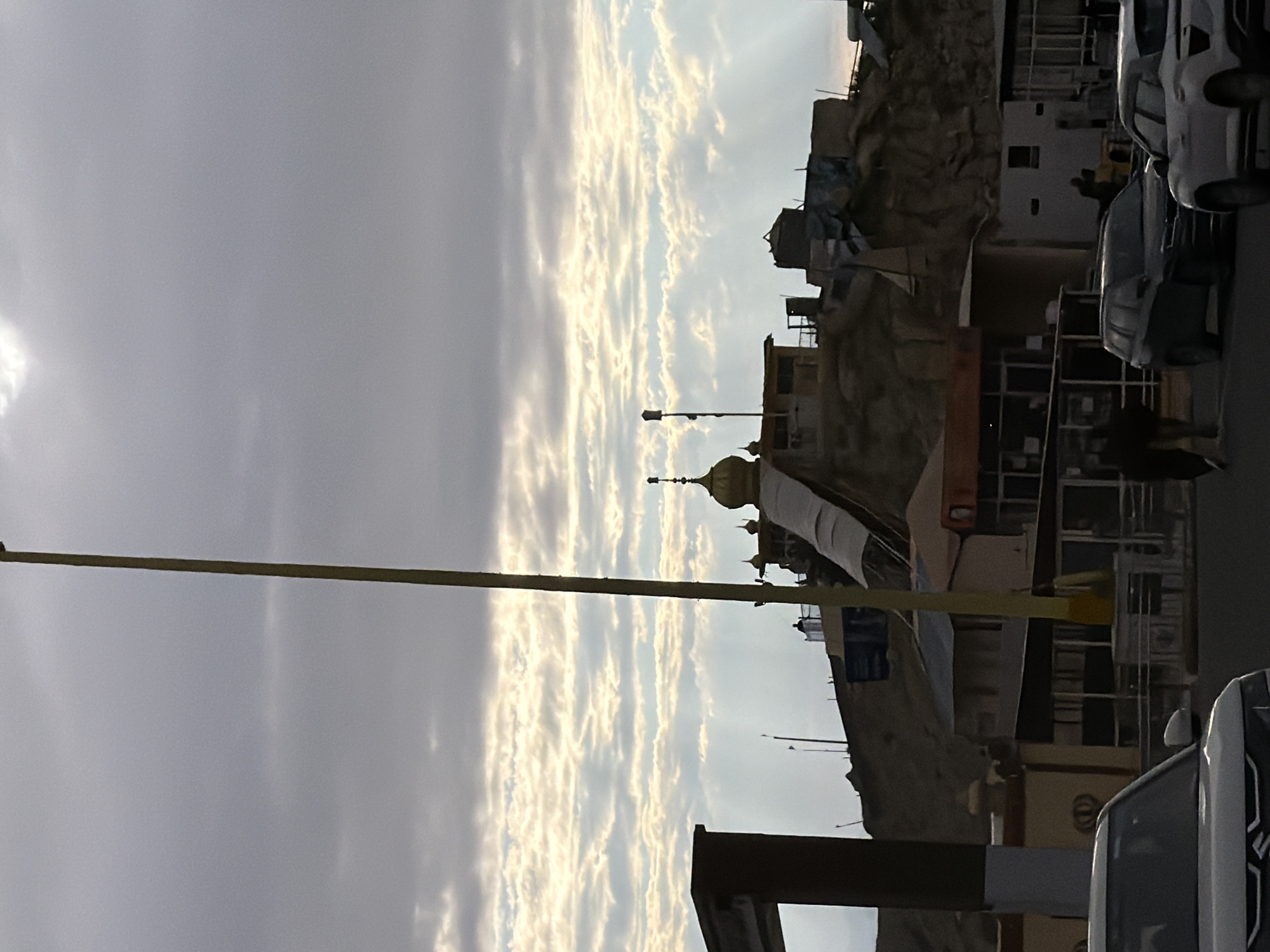 The image depicts a view of the Chumkar Monastery in Chumkar, India, under a cloudy sky. A tall yellow pole stands prominently in the foreground, bisecting the frame vertically. The monastery, situated on a hill, features golden domes and various structures with weathered appearances. Several cars are parked on a street in front of the monastery, and two individuals are walking on the sidewalk. The overall scene is characterized by a slightly overcast sky with visible light rays penetrating through the clouds.