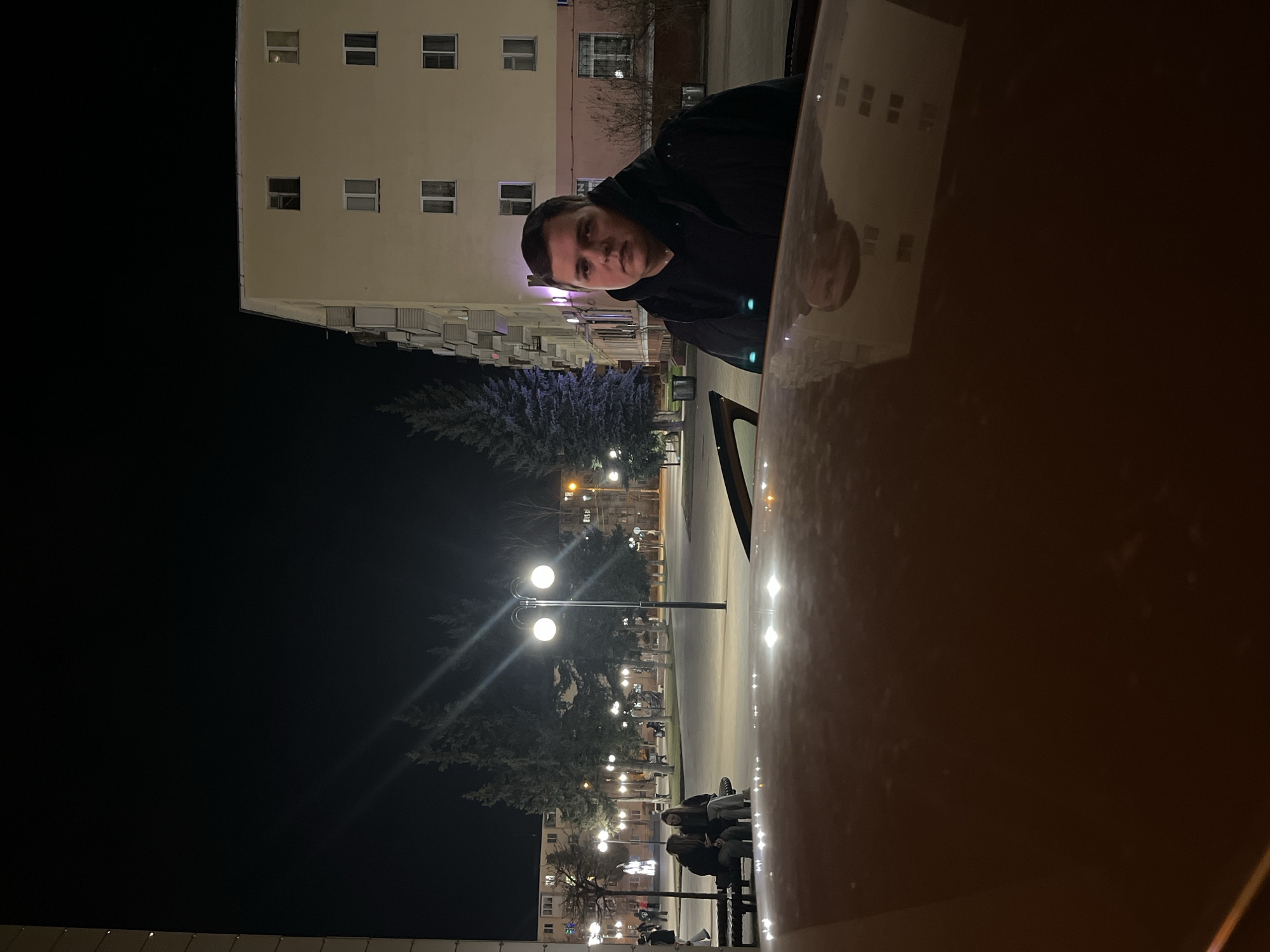 At night, a male is seated in a car in Klin, Russia. The city landscape includes a tall, lit apartment building with multiple windows. Streetlights illuminate a park-like area with trees and benches where people are gathered. The car's hood is visible, reflecting some of the city's lights.