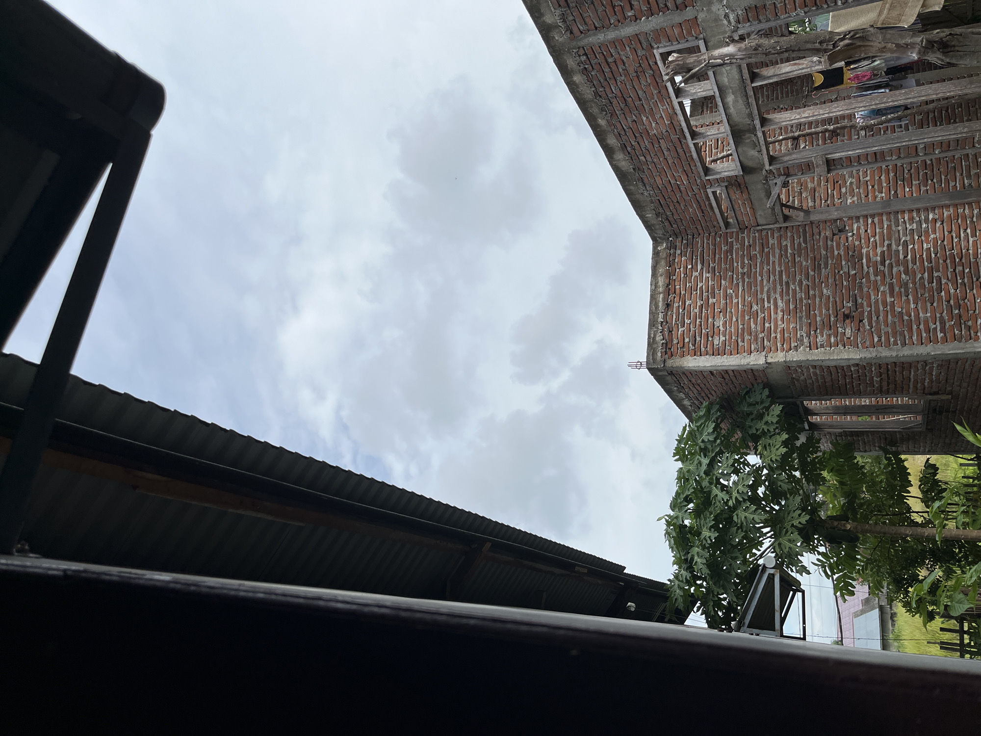 The image depicts an unfinished brick building against a cloudy sky, viewed from beneath the eaves of a corrugated metal roof. To the left, a dark, vertical wooden post frames the scene. In the foreground, a papaya tree with abundant green leaves stands prominently. The building itself is constructed of red bricks with exposed concrete pillars and window frames. Some wooden scaffolding and a clothesline with laundry are visible within the structure. In the background, glimpses of other houses with red roofs and green vegetation suggest a semi-rural or suburban setting within Lhokseumawe, Indonesia. The overall atmosphere is one of ongoing construction and everyday life in a tropical environment.