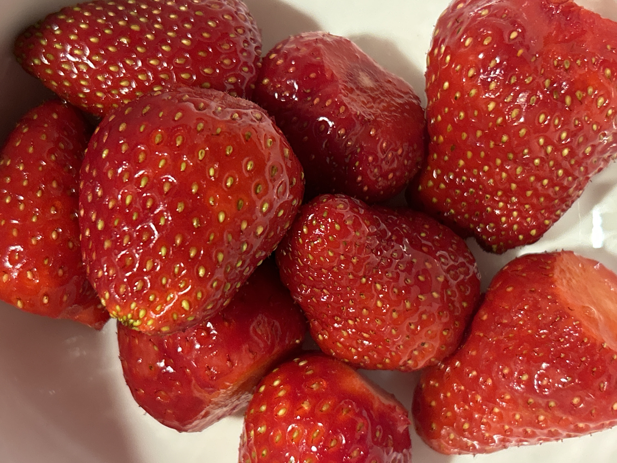The image shows a close-up of a cluster of fresh, ripe strawberries. They are bright red with small yellow seeds on their surfaces. One of the strawberries has a cut edge, revealing the red flesh inside. The strawberries appear to be in a white bowl. No people, actions, or interactions are visible in the image. The location is not specified in the image and there is no basis to assume that they are located in the city of Plovdiv, Bulgaria.