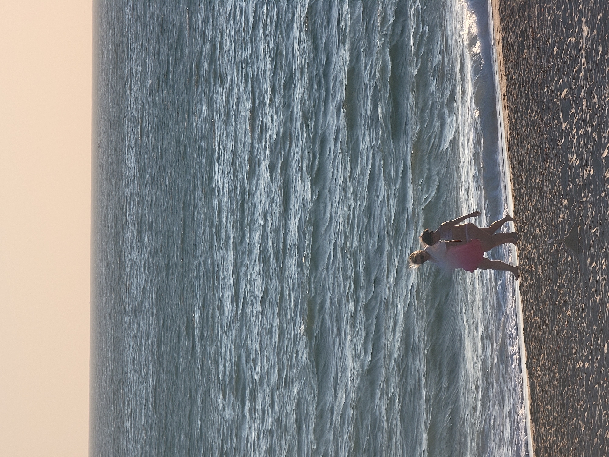 Two individuals, one adult and one child, walk along a sandy beach next to the ocean. The adult, appearing to be female, wears sunglasses and a light-colored top fading into a pink skirt. The child, also appearing female, is dressed in a bikini. They are walking from left to right, parallel to the water's edge. The ocean displays moderate waves and a slightly choppy surface, with a pale, possibly hazy, sky overhead. The beach sand is textured with visible footprints and small indentations. The overall scene captures a leisurely stroll by the sea.