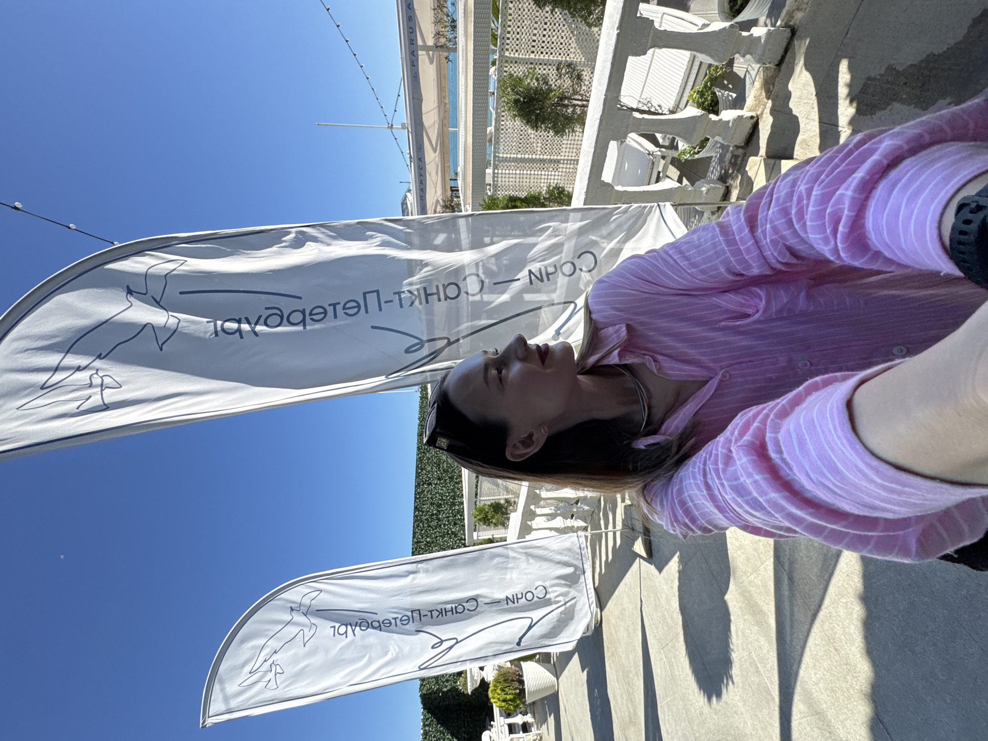 The image captures an outdoor scene on a very bright, sunny day, likely in a coastal resort area. The clear blue sky dominates the upper portion of the frame.
A person is visible on the right side of the photograph, from the chest up, looking upwards and to the left with her face in profile. She has dark hair, wears a pink collared shirt with white stripes, and has sunglasses resting on her head. A dark watch is visible on her left wrist.
Two white flags with a blue design are prominent. The design features a stylized bird in flight. Cyrillic text on the flags reads "СОЧИ — САНКТ-ПЕТЕРБУРГ" (Sochi — Saint Petersburg). These flags suggest a connection or event related to these two Russian cities.
The background reveals white ornate railings and structures, possibly pergolas or buildings with decorative lattice work, typical of a resort or promenade. Green foliage and bushes are visible among the architecture. In the distance, a glimpse of the sea and coastline can be seen. String lights are strung across the top of the frame, indicating a decorated outdoor space. Strong shadows are cast on the paved ground, confirming the bright, direct sunlight. The scene depicts a sunny leisure moment in Sochi, Russia.