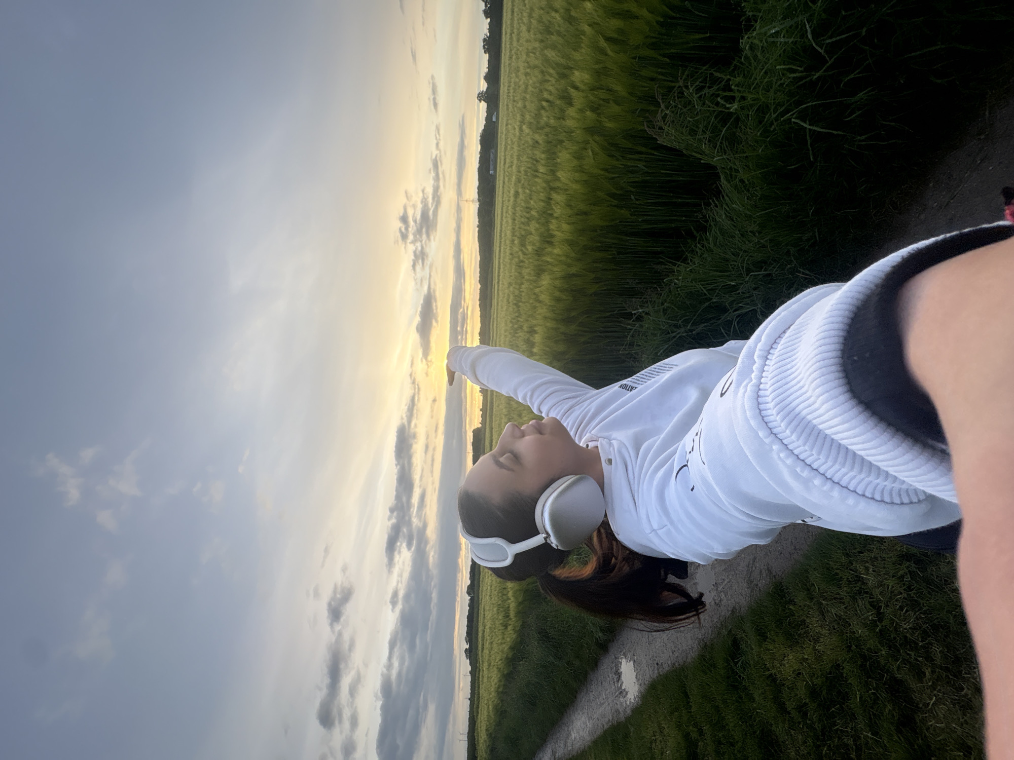 A young person stands on a narrow path next to a field under a sky with clouds. The field is light green and slightly blurred. The sky is blue, with many white and dark gray clouds. A white light shines from the horizon between the field and the sky. The person wears white headphones and a white jacket. Her right arm points toward the light, and her eyes look up. The ground is gray and grainy. The path is bordered by overgrown green grass. The location is Mönchengladbach, Germany.