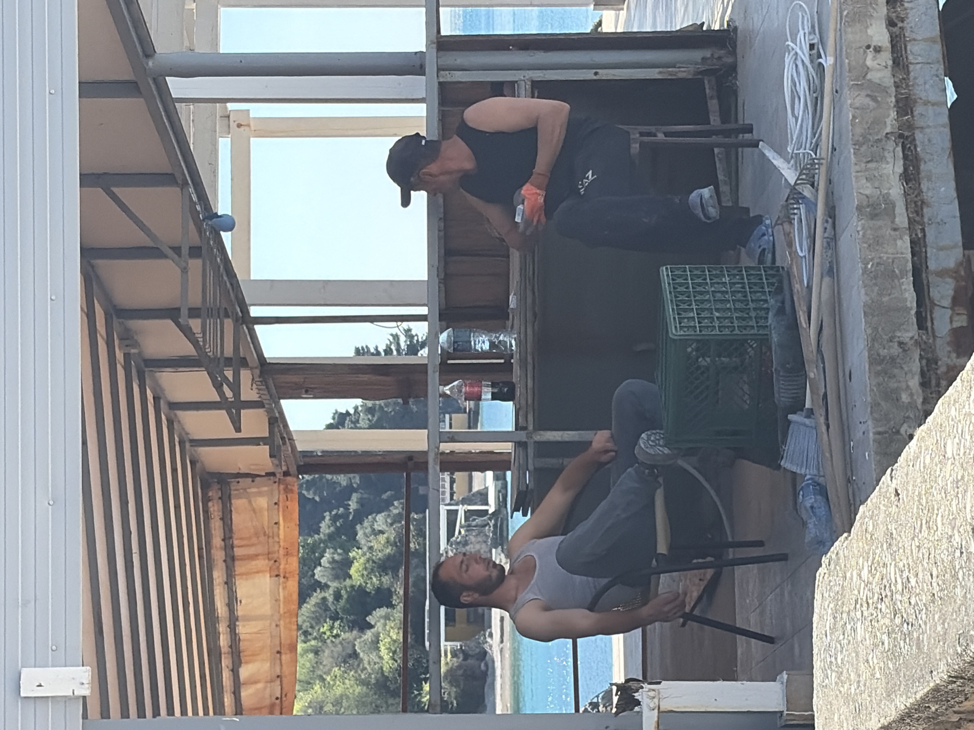 Two men appear to be doing construction work on a building structure. One man is wearing a black cap, black tank top, and dark pants, bending over, as if inspecting or working on something within the structure. The second man is seated in a chair, wearing a grey tank top and jeans, looking downwards and possibly working on something within his reach. The building is open, with the sea and trees visible in the background. There is a Coca-Cola bottle and a water bottle on a horizontal beam. A green crate is next to the seated man. Based on the background landscape, including the sea and vegetation, it is plausible that the location could be in or near Budva, Montenegro.