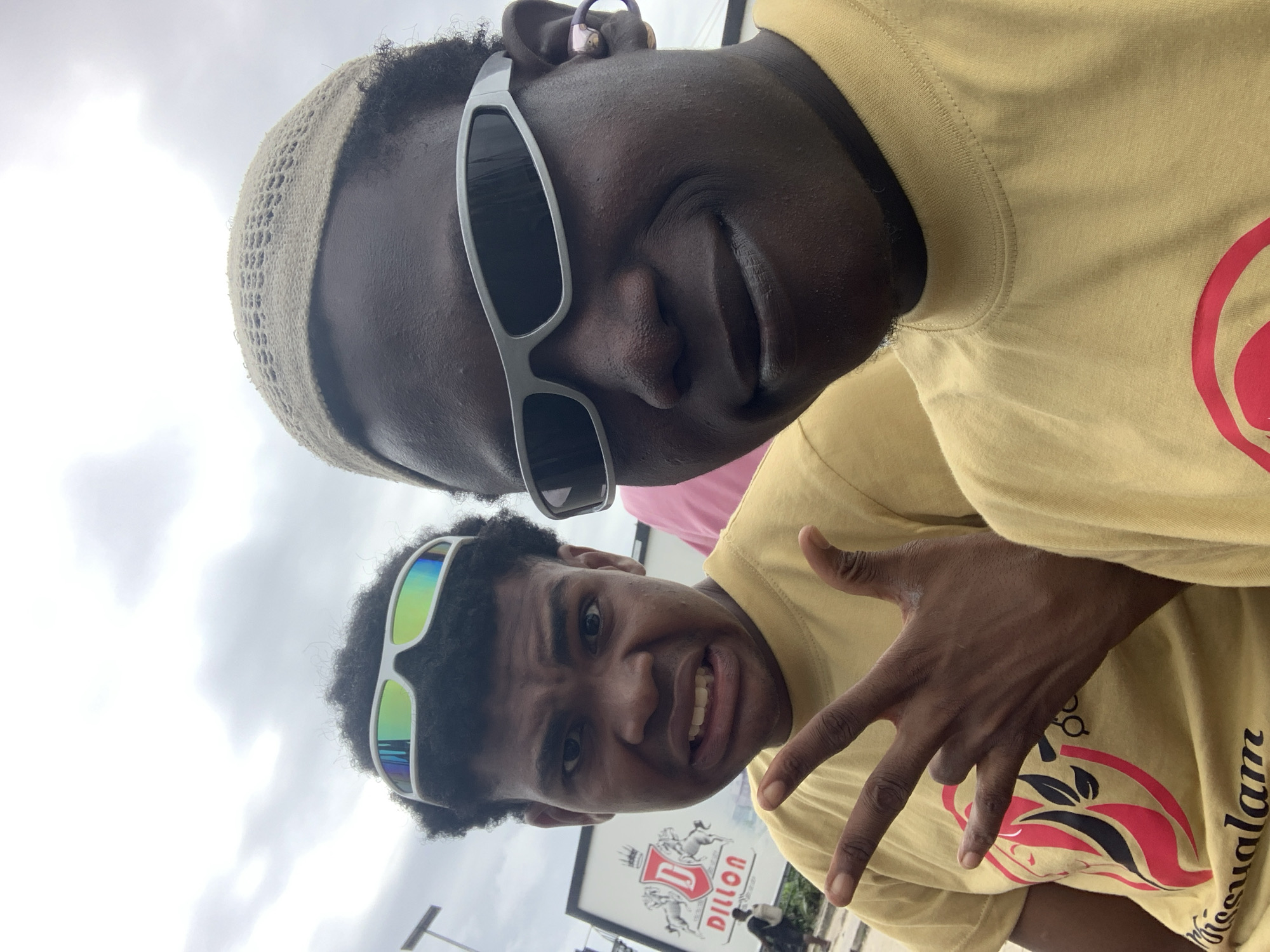Two young Black males are posing for a selfie outdoors. The male in the foreground is wearing a tan knit cap and silver-rimmed sunglasses. He is looking directly at the camera with a slight smile. A pair of purple earbuds is visible in his right ear. He is wearing a yellow t-shirt with a graphic design and text that reads "visualam" at the bottom. The male behind him is wearing white-rimmed, multi-colored reflective sunglasses and a yellow t-shirt. He is making a grimace and holding up three fingers on his left hand in a hand gesture. In the background, a large white billboard with the "DILLON" logo and two horses is visible. The sky is overcast with clouds. The location is identified as the city of Ilado, Nigeria.