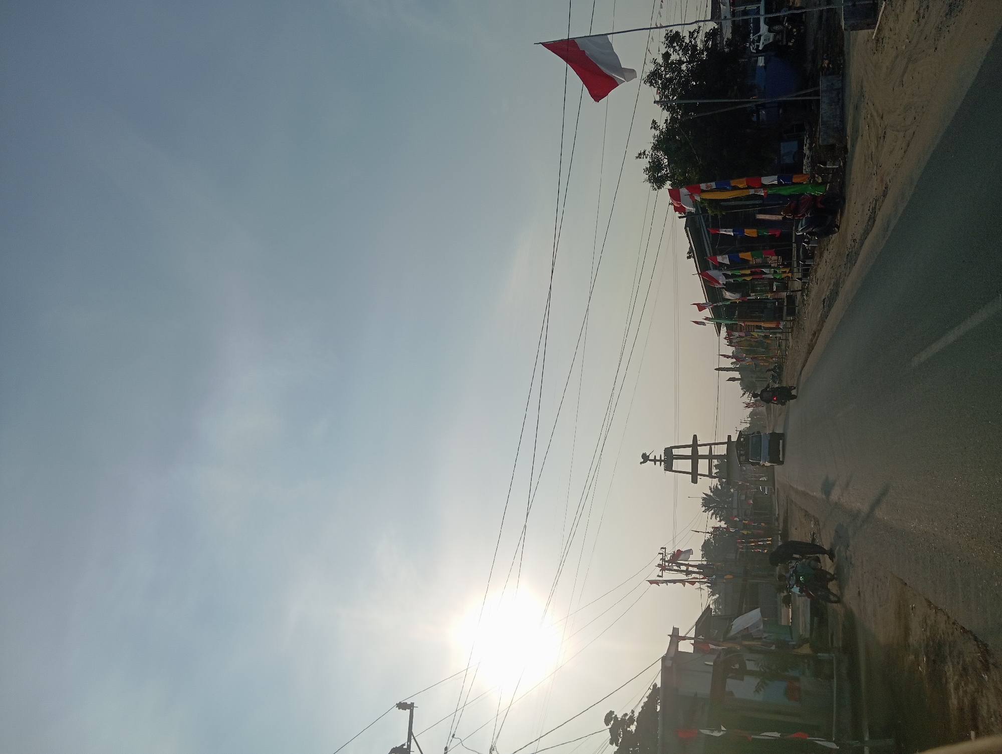 The image depicts a street scene in Dente Makmur, Indonesia, under a bright, hazy sky. The sun is low in the sky, casting a bright glare and creating a soft light. The street is paved and appears to be a two-lane road. On both sides of the road are buildings, some appearing to be shops or homes, with various flags and decorations displayed. The flags are predominantly red and white, suggesting a national celebration or holiday.
On the left side of the road, there are several small buildings and a utility pole with multiple wires. A motorcycle is parked on the dirt shoulder, and a person is standing next to it.
On the right side of the road, there is a row of shops or businesses, also adorned with numerous flags. A motorcycle is parked in front of one of these buildings. Further down the road, there is a larger, darker building with trees behind it. A white SUV is parked in front of this building.
In the distance, down the center of the road, a tall, multi-tiered structure topped with a figure resembling a rooster is visible. A van is driving towards this structure. Another motorcycle is seen on the right side of the road, further down. The overall impression is of a populated area, possibly a village or small town, decorated for a special occasion. Power lines crisscross the sky throughout the scene.