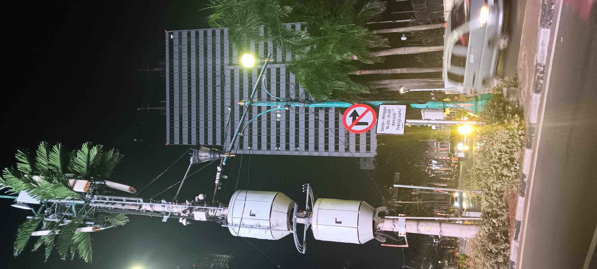The image depicts a night scene in Central Jakarta, Indonesia, near a bus rapid transit station. In the foreground, a road curves to the right with a concrete median adorned with green foliage. A light-colored van is captured in motion, its headlights illuminated. Behind the road, a tall, modern building with a grid-like facade dominates the background. To the left, a utility pole, designed to resemble a palm tree, supports several antennas and surveillance cameras. A street lamp with a decorative green pole stands near the building. A traffic sign, indicating "No Right Turn" with a time restriction of "Senin - Minggu 16.00 - 21.00" (Monday - Sunday 4:00 PM - 9:00 PM) and an exception for "Kecuali TransJakarta" (Except TransJakarta), is prominently displayed. The surrounding environment is illuminated by artificial lights, casting a warm glow on the scene.