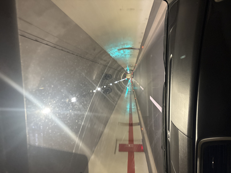 The image captures a forward-facing view from inside a vehicle traversing a two-lane road tunnel in Donje Mrke, Montenegro. The tunnel features an arched, light-colored concrete ceiling with overhead white lighting and multiple circular ventilation fans. The walls are predominantly light-colored. The left tunnel wall presents a red horizontal stripe intersecting with a vertical red stripe, forming a cross pattern, adjacent to a red emergency door. The right tunnel wall contains segments illuminated by blue-green lights and an observable alcove further down the tunnel. The roadway is dark asphalt with white solid and dashed lane markings. In the far distance, a white truck with illuminated red taillights is visible. The lower portion of the image shows the vehicle's black dashboard. Reflections of the tunnel lights are present on the windshield, which also bears a faint digital overlay "H3.702.026" in the upper left corner.