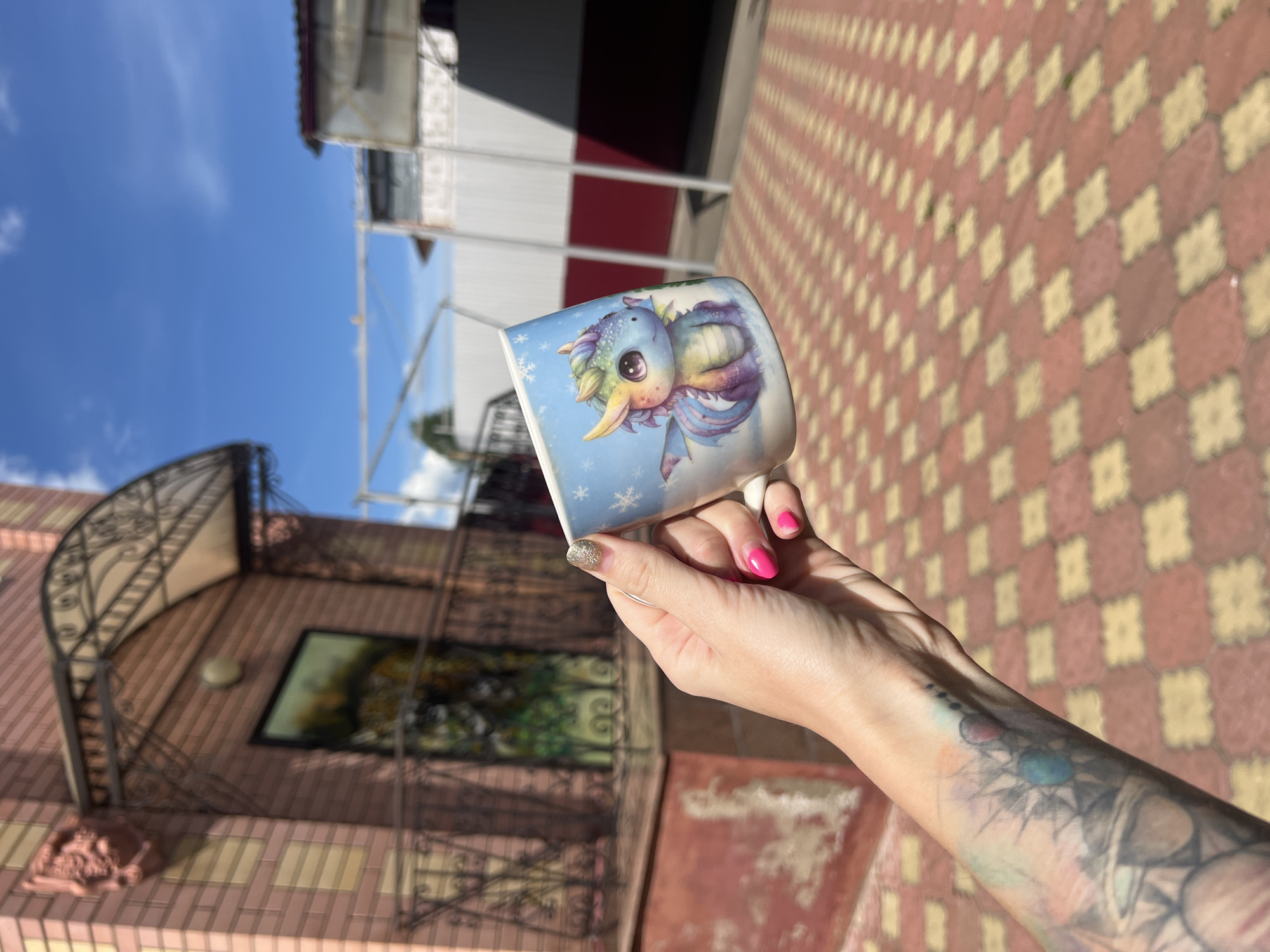 A person's hand, adorned with a galaxy-themed tattoo on the forearm and bright pink nail polish, holds a white ceramic mug. The mug features a colorful cartoon dragon illustration against a light blue, snowflake-patterned background. The person is standing outdoors on a red and yellow checkered paved area, with a light-pink brick building and a wrought-iron fence visible behind them. The scene is under a clear blue sky with scattered white clouds. The background also shows a white fence with red trim and some scaffolding. The location is identified as Krasnoe-na-Volge, Russia.