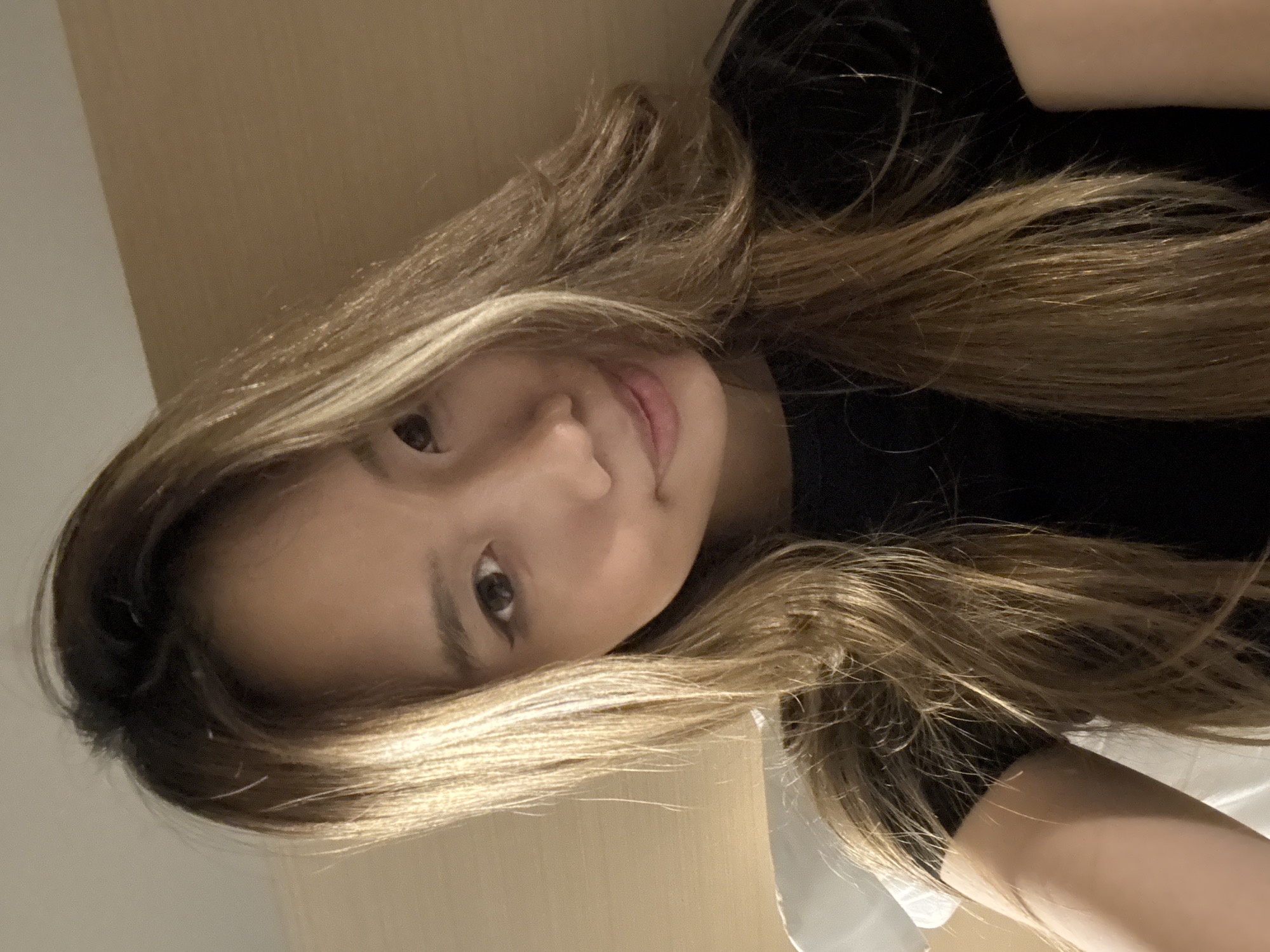 A young East Asian woman with shoulder-length, layered, blonde-brown hair, wearing a black t-shirt, is shown in a close-up selfie. Her head is tilted slightly to her left, and she is looking directly at the camera. She has light brown eyes and is wearing subtle eye makeup. The background appears to be a plain, light brown wall, possibly in a hotel room. The lighting is warm and casts a slight shadow on the right side of her face. The image was taken in Yuzhong District, China.