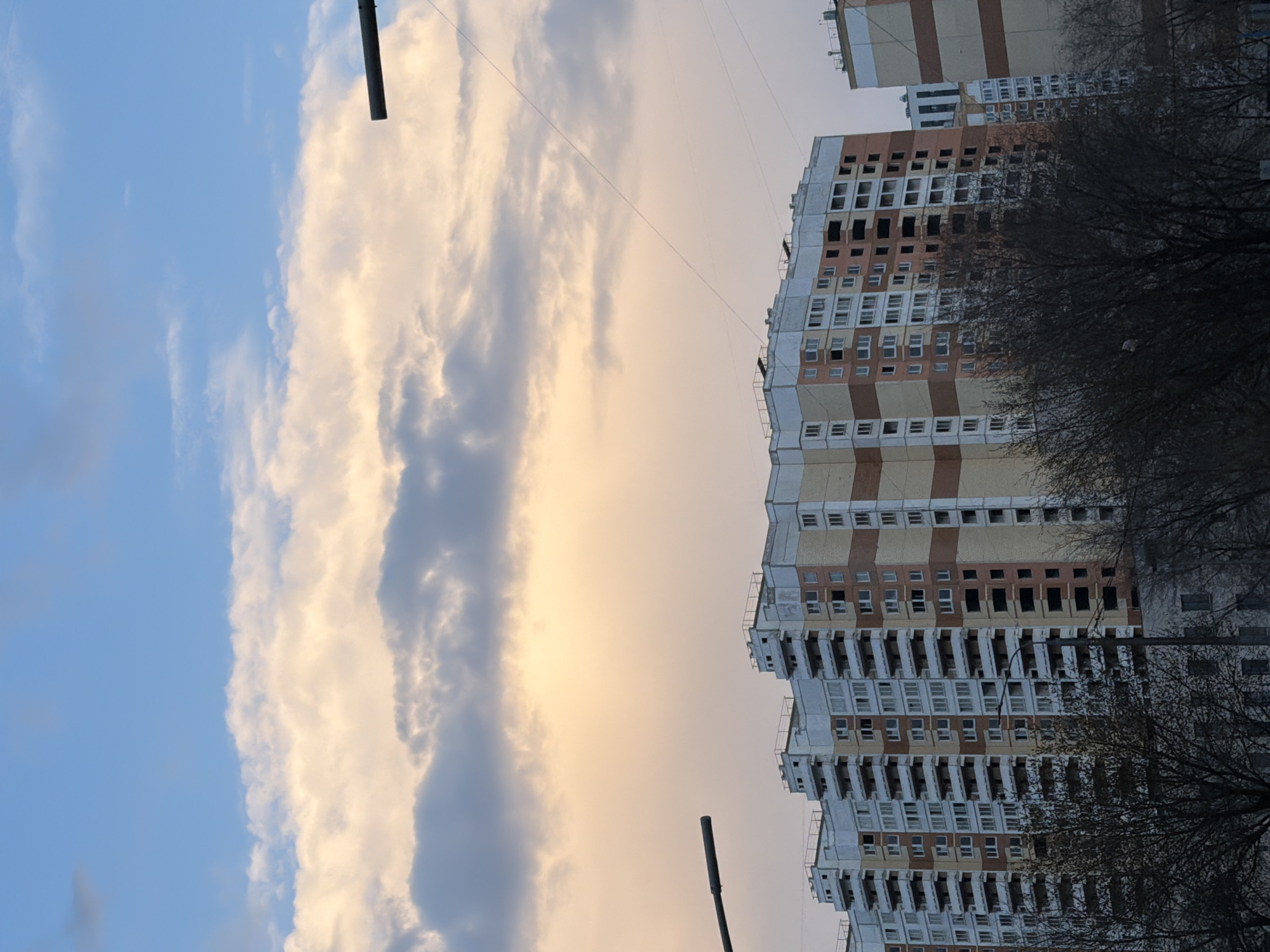 The image shows a multi-story residential building in Moscow, Russia, under a sky with clouds and pale sunlight. The building has a light facade with brown and white horizontal stripes. Bare branches of trees are visible in front of the building. Electrical wires stretch across the sky. The lighting suggests either sunrise or sunset.