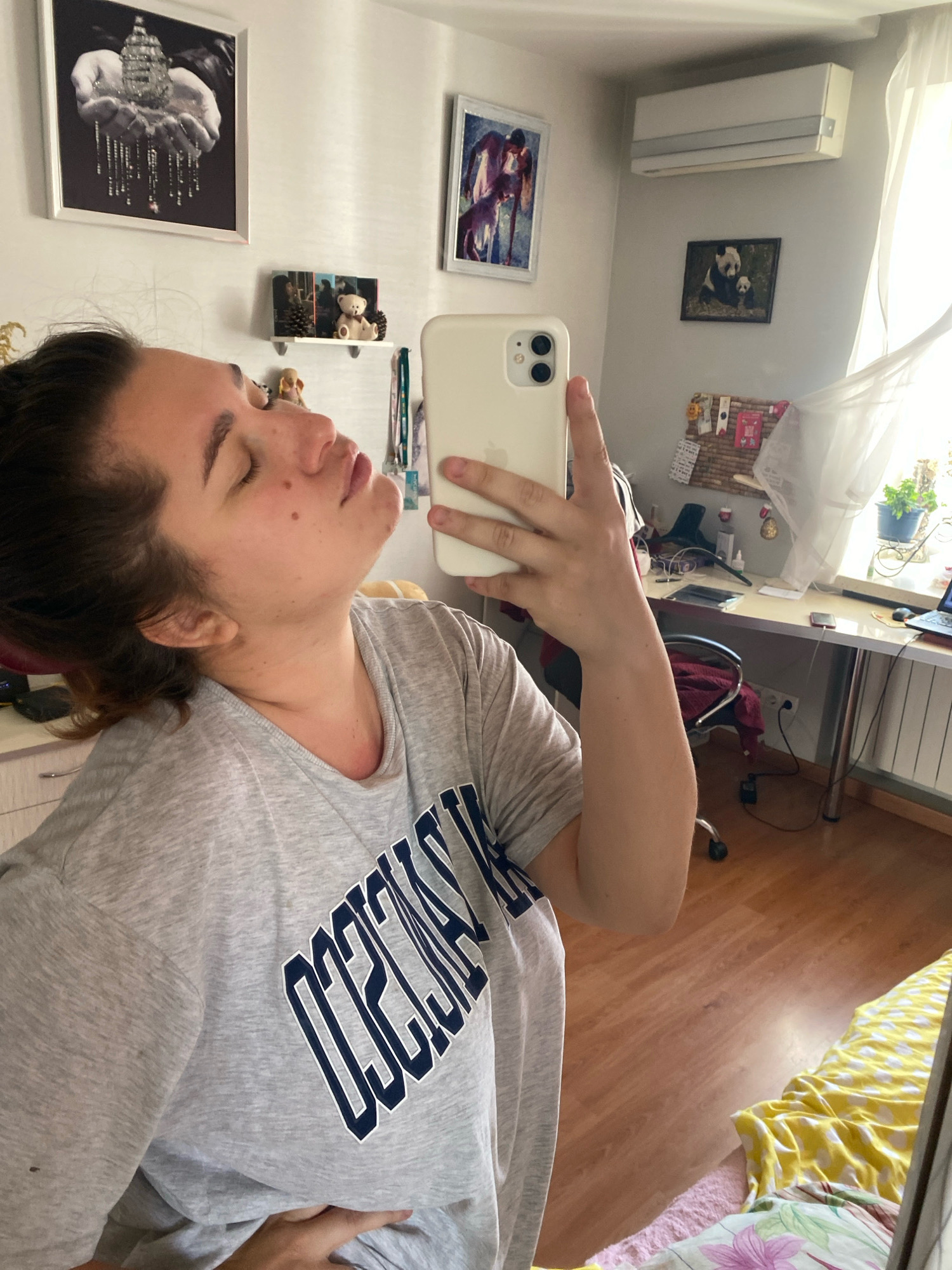The image depicts a young woman taking a selfie in what appears to be her bedroom. She is wearing a grey t-shirt with blue lettering. Her hair is dark and pulled back. She is looking at her phone, which she is holding in her right hand. The room has a desk with a laptop and other items on it, a chair, and several framed pictures on the wall. A window with sheer curtains is visible on the right side of the image. The floor is wooden. Based on the available visual information, there is no definitive indication of the city of Perm, Russia.