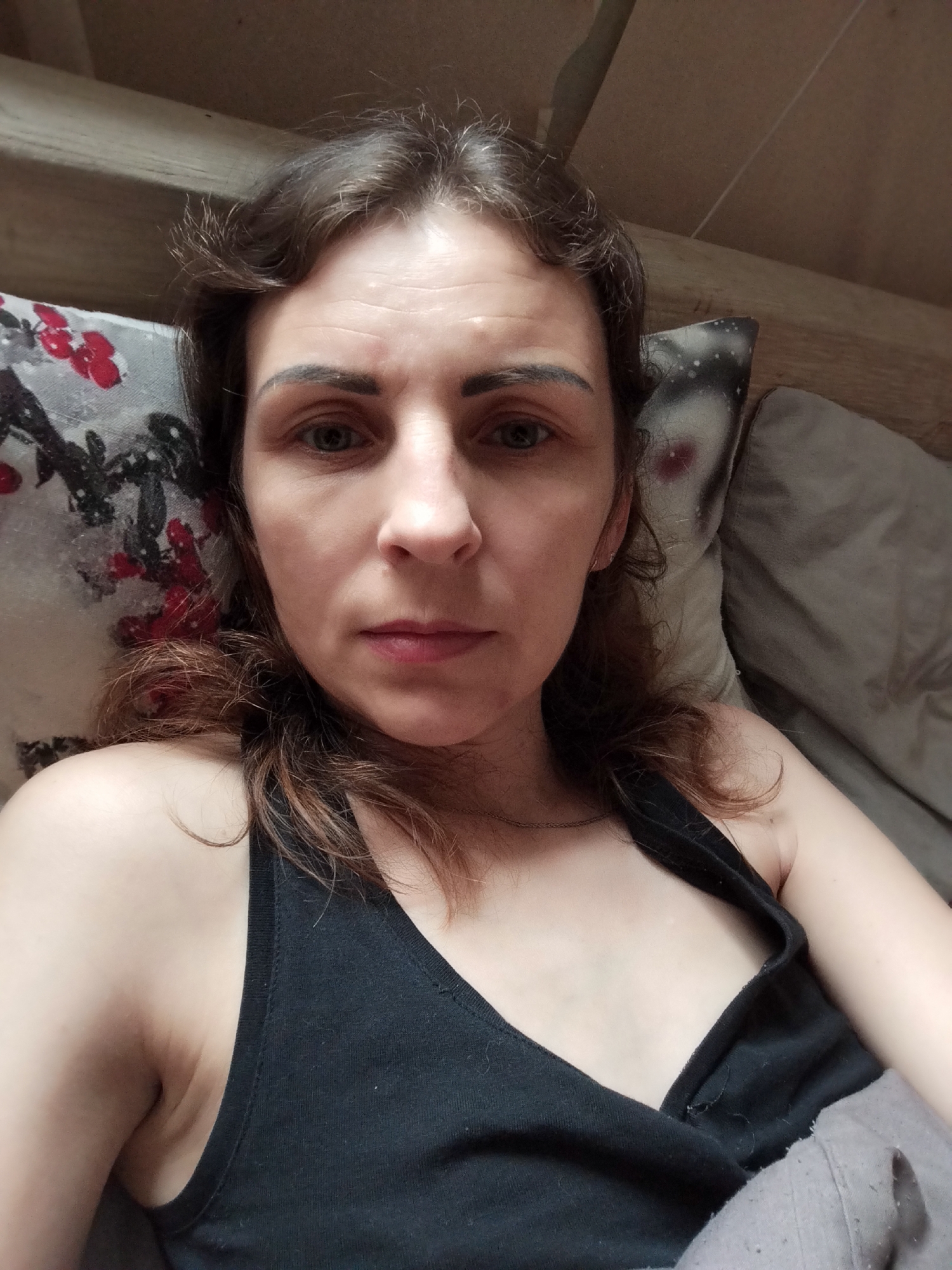 The image shows a woman lying down, possibly in bed, with her head resting on a pillow. She is looking directly at the camera. Her hair is brown and slightly messy. She is wearing a black tank top. The background appears to be a wooden wall and a pillow with a floral pattern. There is no indication of the location being in Vinnytsia, Ukraine.