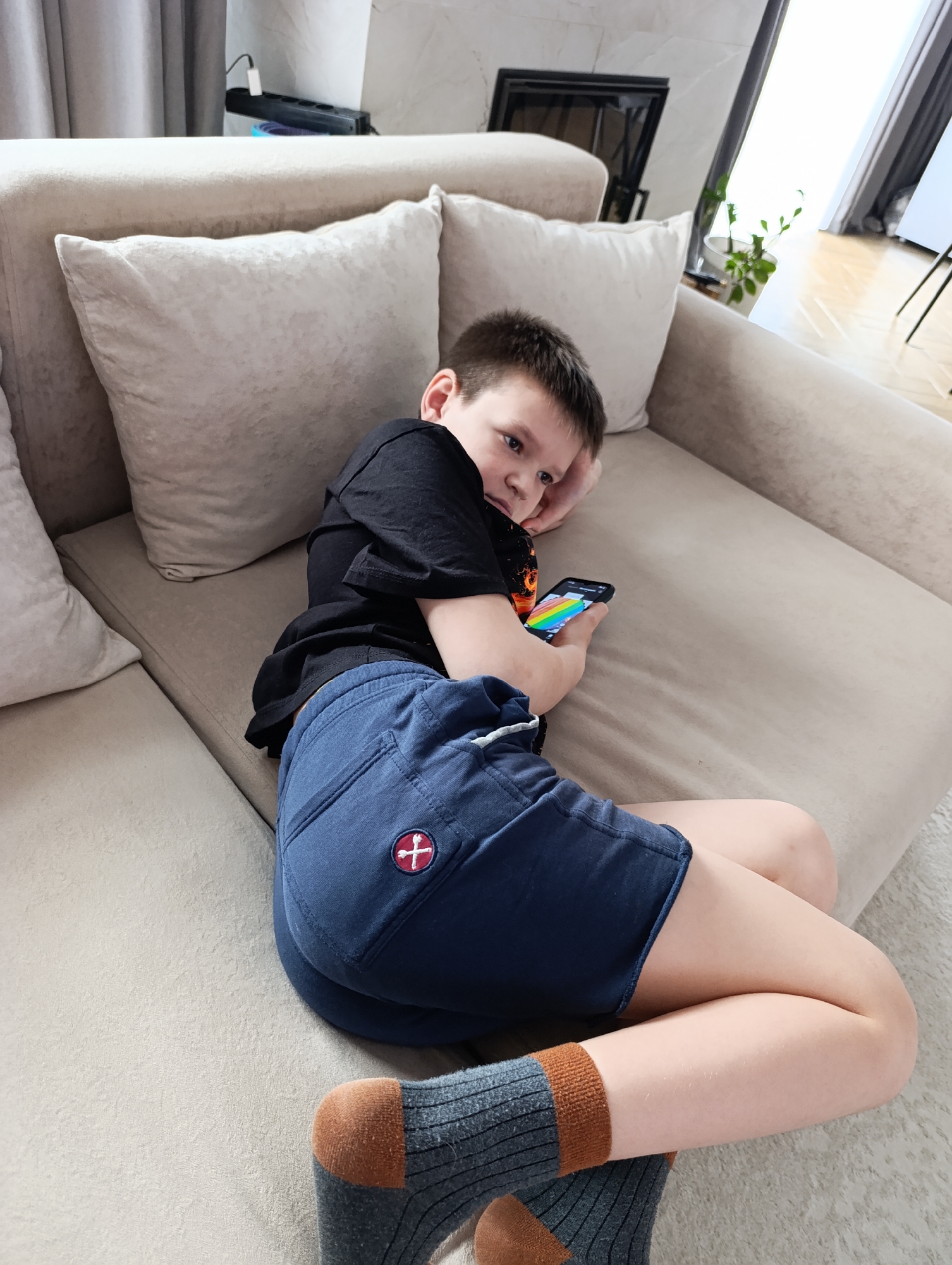 A young boy is captured in a relaxed, candid pose, lying on a light-colored fabric sofa indoors in Chaikovsky, Russia. He is positioned on his side, propped up on one elbow, and appears to be engrossed in his smartphone which he holds in his left hand. The phone screen displays a vibrant, colorful image resembling a rainbow.
The boy has short, dark hair and is wearing a black t-shirt, navy blue shorts with a distinctive red and white cross-like logo on the right thigh pocket, and grey socks with brown heels and toes. His posture suggests leisure and comfort.
The setting is a modern living space, indicated by the contemporary design of the sofa with its plush throw pillows. In the background, to the left, light grey curtains are partially drawn, and a black power strip with a white plug can be seen on a surface. To the right, through a large window or glass door, bright natural light streams in, illuminating a wooden floor and a small potted plant. A dark-framed object, possibly a decorative fireplace insert or wall art, is also visible near the window. The overall ambiance is bright and casual, typical of a daytime scene in a residential home. There is no other discernible text visible in the image besides the logo on the shorts and the image on the phone screen.