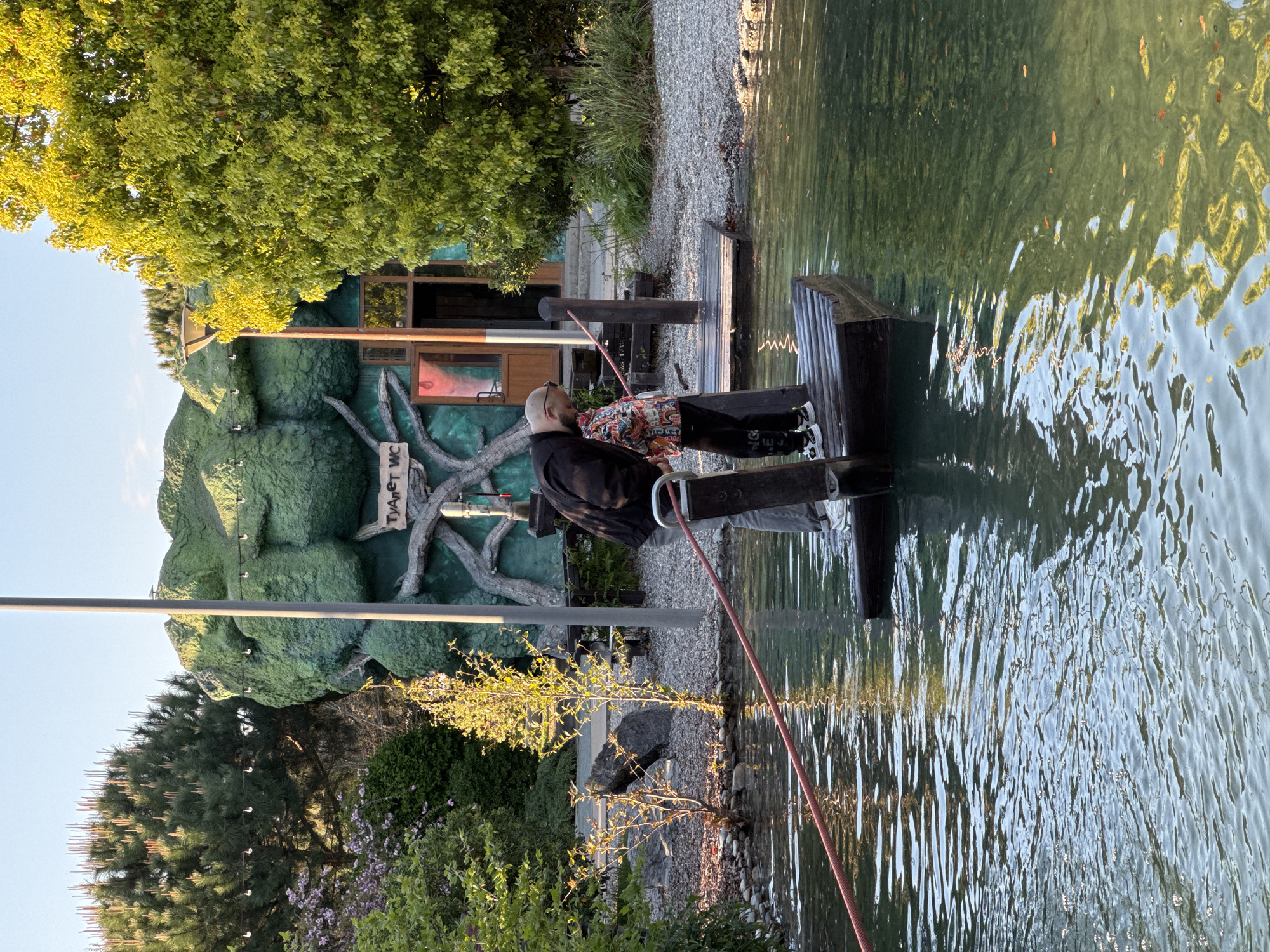 Here is a detailed description of the image:
**Overview:** The image depicts a small water feature or pond in what appears to be an outdoor public area. Two individuals are present, positioned on a wooden structure extending over the water. The setting includes landscaped elements such as trees, shrubs, and gravel pathways. A distinctive building or decorative structure with a tree-like facade is visible in the background.
**People:**
* Two individuals are standing on the wooden structure. One is wearing a black shirt and jeans, leaning down with a red pole or hose extended towards the water. The other individual is wearing a patterned jacket and black pants, standing behind the first person.
**Subjects and Objects:**
* **Water Feature:** A pond or water feature with rippled water surface.
* **Wooden Structure:** A wooden platform or dock extending into the water.
* **Red Pole:** A long, red pole or hose, possibly used for some activity in the water.
* **Tree-like Facade:** A building or decorative structure with a unique facade resembling a large, green tree or mountain. There is signage that reads 'Туалет WC'.
* **Landscaping:** Trees, shrubs, gravel, and other landscaping elements.
**Actions and Interactions:**
* The two individuals are positioned on the wooden structure near the water.
* One of the individuals is holding a red pole or hose and interacting with the water.
**Environment:**
* **Location:** An outdoor area with landscaping and a water feature. Given the signage written in Russian, it's likely located in a Russian-speaking area.
* **Weather/Time:** The lighting suggests either late afternoon or early morning. The weather appears to be clear and sunny.
**Location in Sochi, Russia:**
* The signage 'Туалет WC' indicates that the structure is the Russian word for toilet with an added WC (water closet). Therefore, the building in the background is a unique toilet area design.
* Given the decorative tree-like structure and location details, it is likely located in Lazarevskoye Microdistrict, Sochi, Russia.