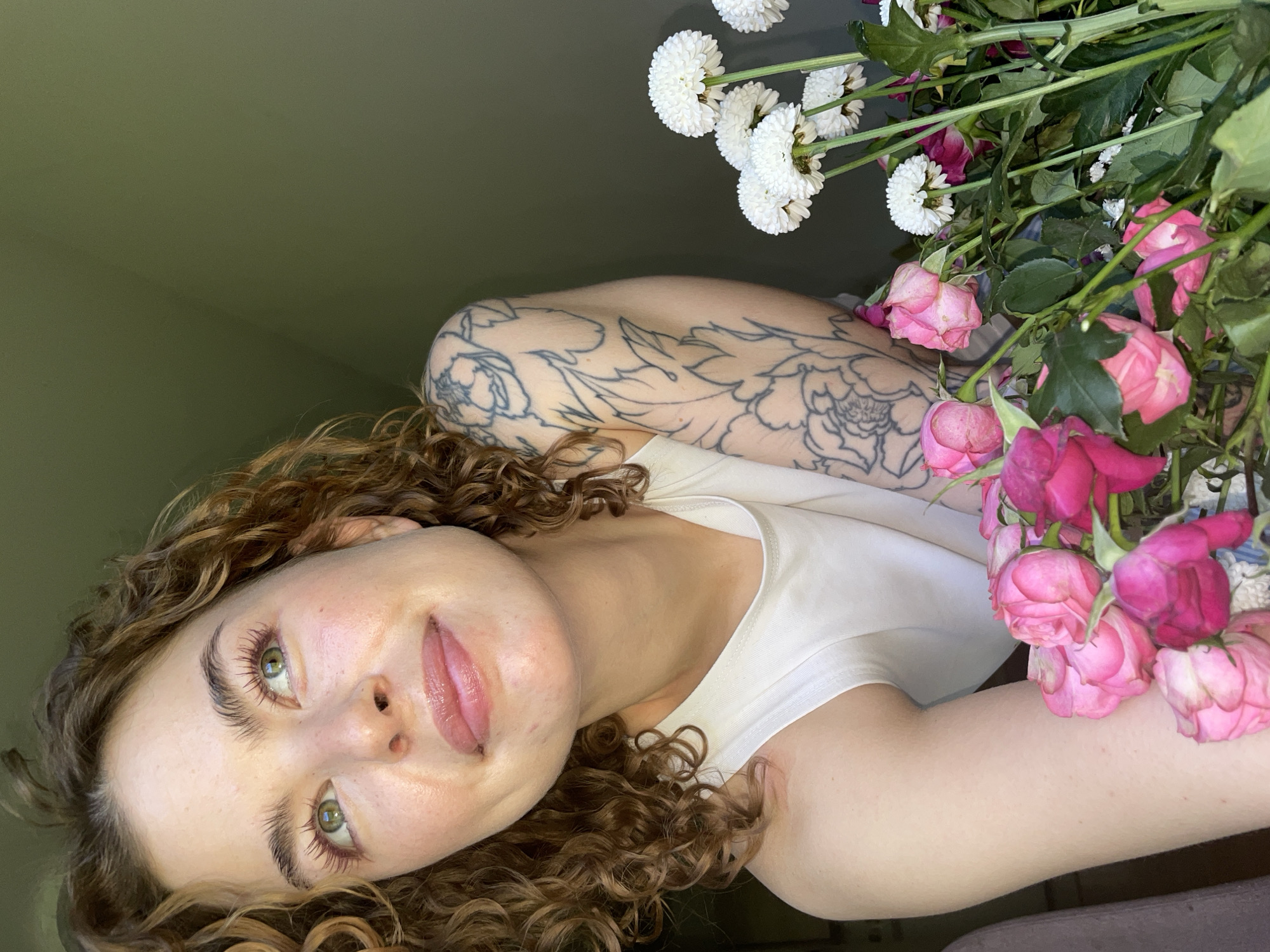 A medium close-up shot, oriented vertically, features a young woman posing with a bouquet of flowers against a plain olive-green wall. The woman has long, curly brown hair that frames her face, green eyes, and a light complexion, looking directly at the camera with a subtle, neutral expression. Her lips are slightly parted, revealing a hint of a smile. She is wearing a simple white tank top. A prominent, uncolored tattoo of floral or botanical patterns covers her left upper arm and shoulder.
A mixed bouquet of flowers is positioned to the right of the woman, with some overlapping her right shoulder and arm. The bouquet contains white, spherical chrysanthemums and several pink roses, some of which appear slightly wilted or past their prime. The plain background suggests an indoor setting. There is no visible text. The provided location context for this image is Boreti, Montenegro.