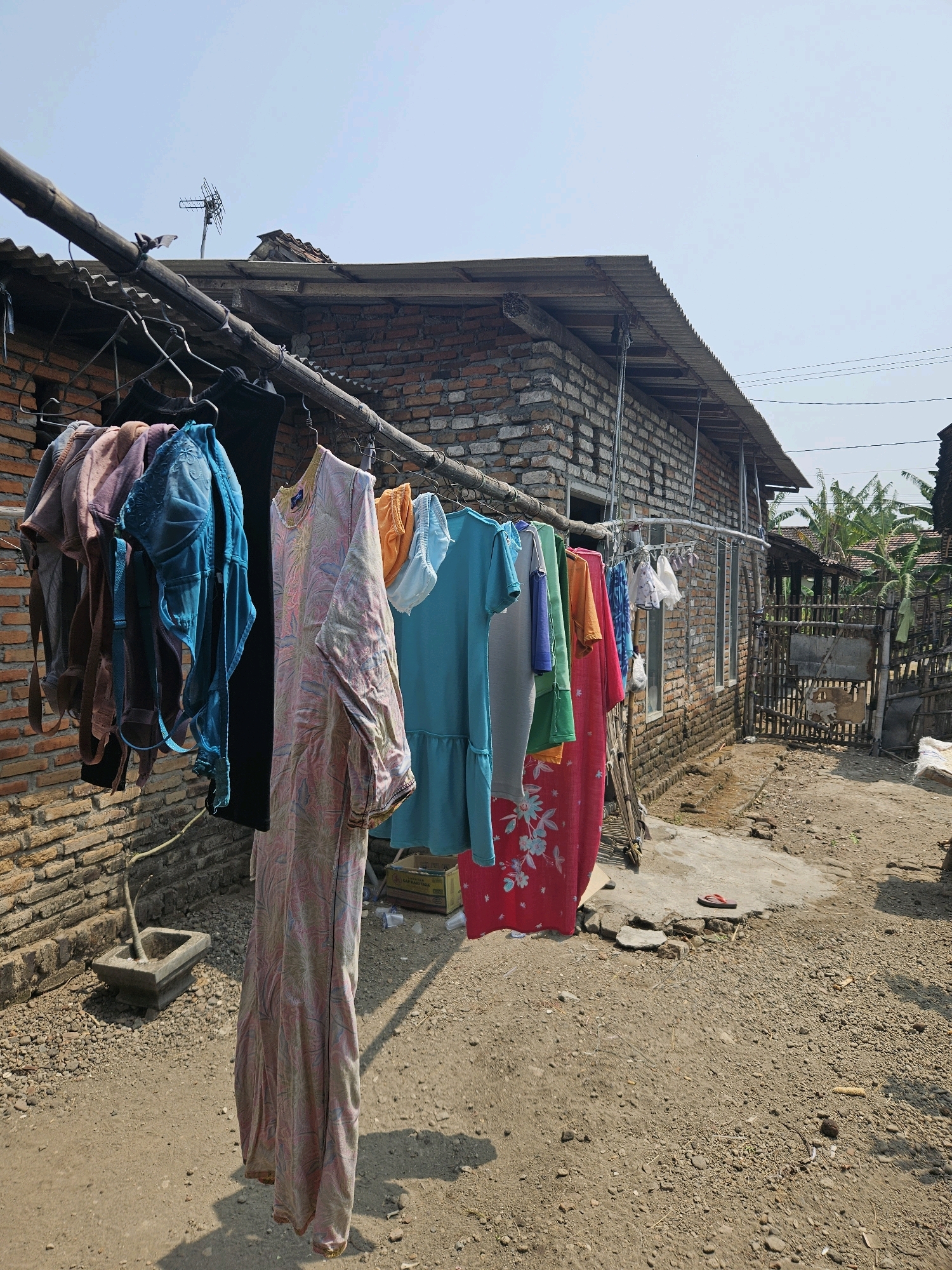 A sunny daytime, outdoor scene in Kampungbaru, Indonesia, shows a brick building with a corrugated metal roof. Laundry hangs on a clothesline strung between poles in front of the building. The laundry includes several bras, a long patterned dress, a bright blue dress, and other garments. The ground is covered in gravel and dirt. A wooden fence and some banana trees are visible in the background. The scene depicts a typical residential setting.