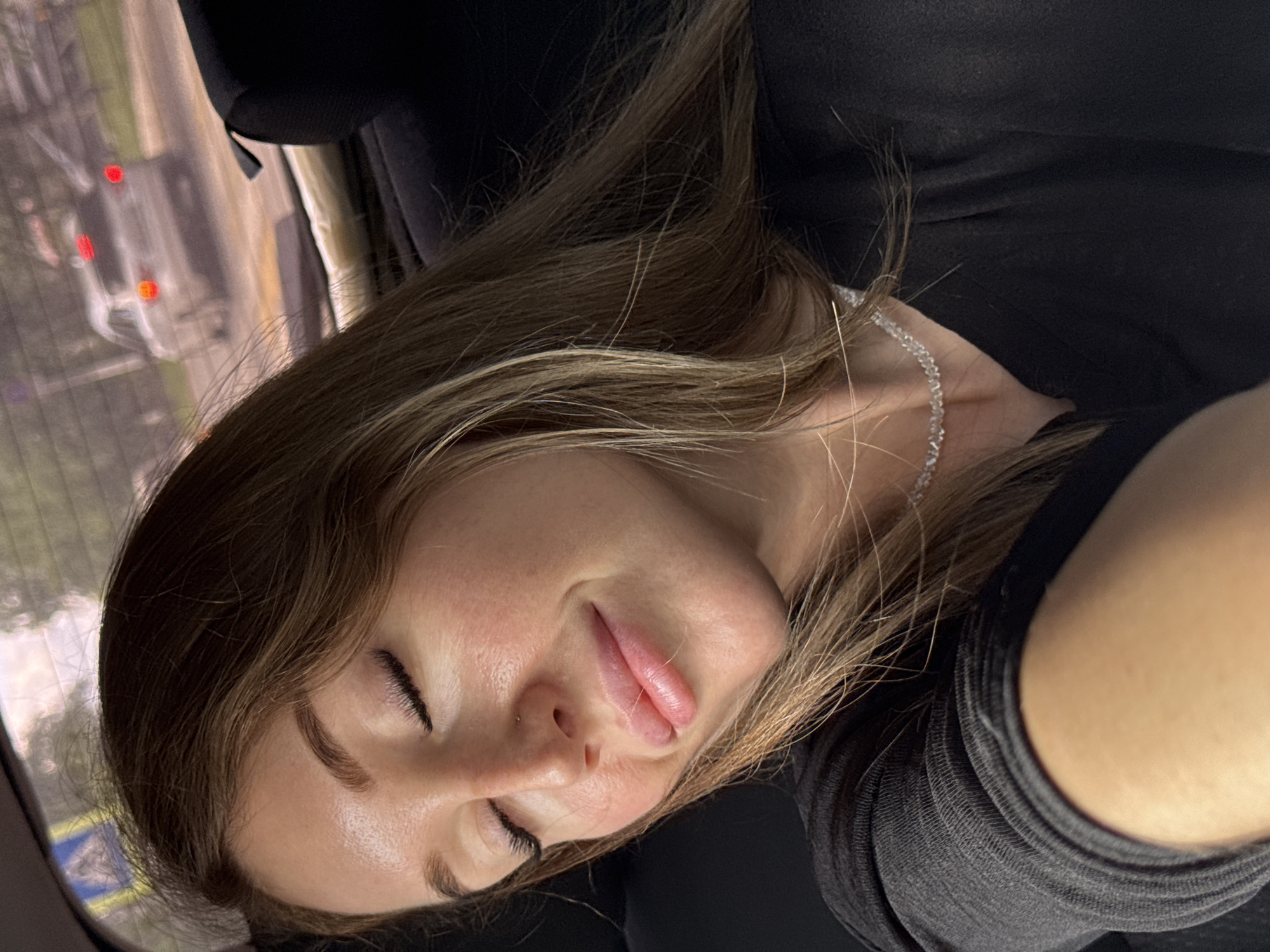 A young woman with long brown hair is resting with her eyes closed and a slight smile on her face in the back seat of a car. She is wearing a black shirt and a thin, sparkly necklace. The background shows a blurry view through the car window, with traffic and trees visible. There are no specific identifiable landmarks that would pinpoint the location to Chaikovsky, Russia.