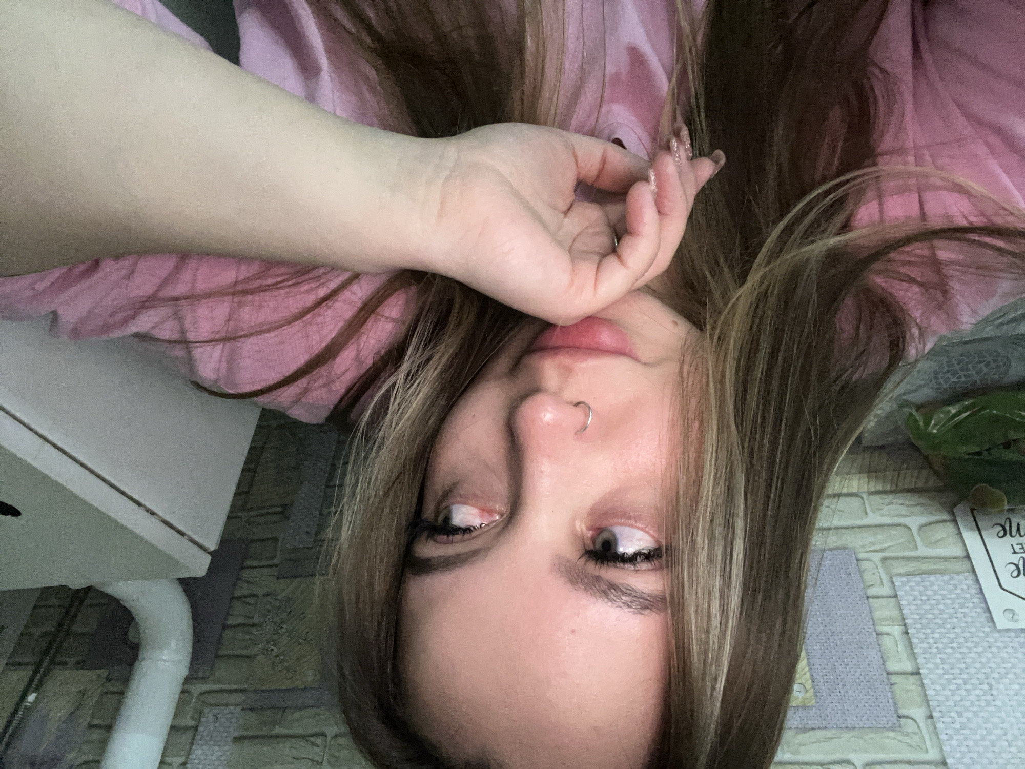 A young woman is depicted in a close-up, inverted photograph. She has light skin, brown hair with blonde highlights, and a nose piercing with a silver ring. Her left hand is raised, with fingers near her mouth. She wears a pink top.
The background includes part of what appears to be a white desk or cabinet, along with a section of a wall or surface with a brick-like pattern. A small, partially visible object with text is in the background, slightly blurred.
There is no indication of Port-Katon, Russia.