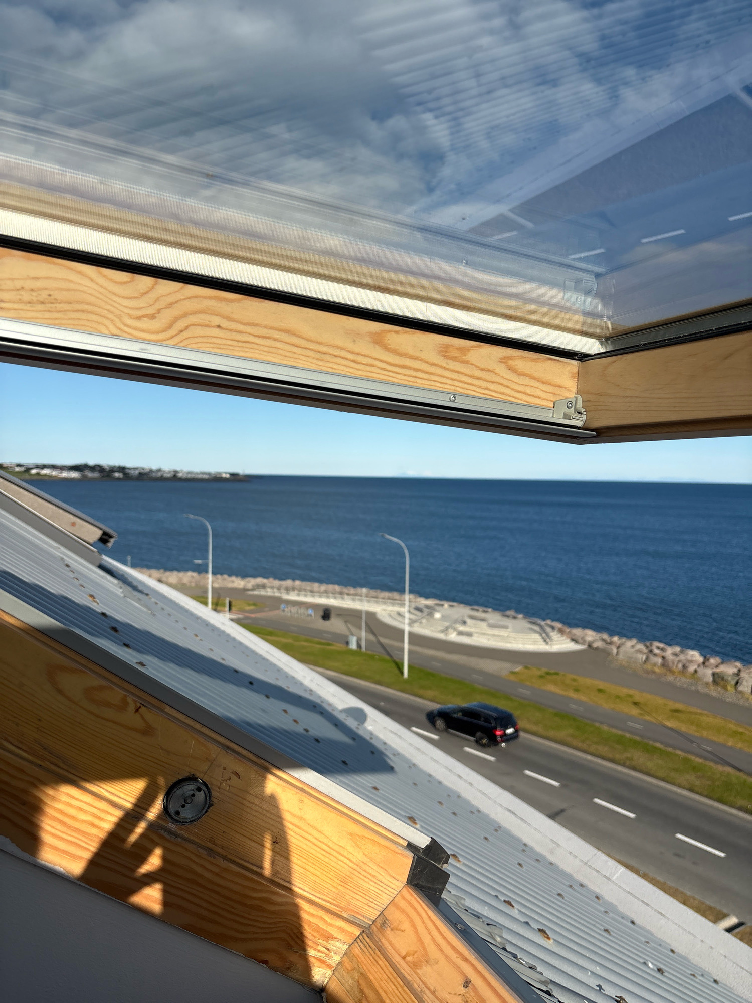 An eye-level, close-up, vertical, exterior shot captures a view through an open skylight towards the sea and a coastal road in Reykjavik, Iceland. The skylight's wooden frame, with visible grain and some discoloration, occupies the foreground and a significant portion of the top of the frame. A metallic track and hinge mechanism is visible along the edge of the opened skylight. Below the skylight, a corrugated metal roof slopes downwards.
Beyond the roof and skylight, a wide expanse of deep blue sea stretches to the horizon, meeting a clear, bright blue sky with scattered, wispy white clouds. Along the coastline, a paved road is visible, with white dashed lines indicating lanes. A black SUV is in motion on this road, heading towards the left of the frame. Adjacent to the road, a grassy area and a curved concrete structure, possibly a walkway or viewing platform, are present. In the far distance, a low-lying coastline with buildings and a lighthouse can be discerned. The overall scene is bright and sunlit, suggesting a clear day.