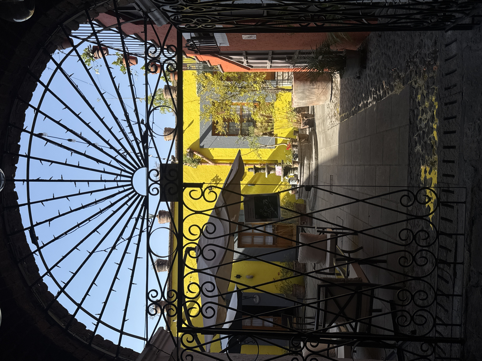 A vibrant, sunlit courtyard in Puebla, Mexico, is framed and viewed through a dark, ornate wrought-iron gate. The gate, occupying the foreground, features an elaborate semi-circular fan design on the left, adorned with small spikes or string lights, and intricate scrollwork on the right.
Beyond the gate, a brightly painted yellow building with a contrasting gray lower section and a red-orange roofline stands out. Wooden-framed windows and a partially unfurled white umbrella or canopy are visible on the building. Green foliage, possibly a small tree or bush, grows near the building, adding a natural element to the colorful facade. Several potted plants hang suspended against the clear blue sky, visible through the upper sections of the gate.
To the right, a dark, textured stone wall runs alongside a paved path or walkway, with patches of yellow paint or decorative accents on its surface and the ground. The strong, direct sunlight casts clear shadows, indicating it is daytime, likely morning or afternoon, under a clear sky.
No people are visible in the scene. A dark sign with an emblem or text, too indistinct to read, is affixed to the yellow building. The architectural style, vibrant colors, and sunny disposition evoke the typical charm of a Mexican colonial city.