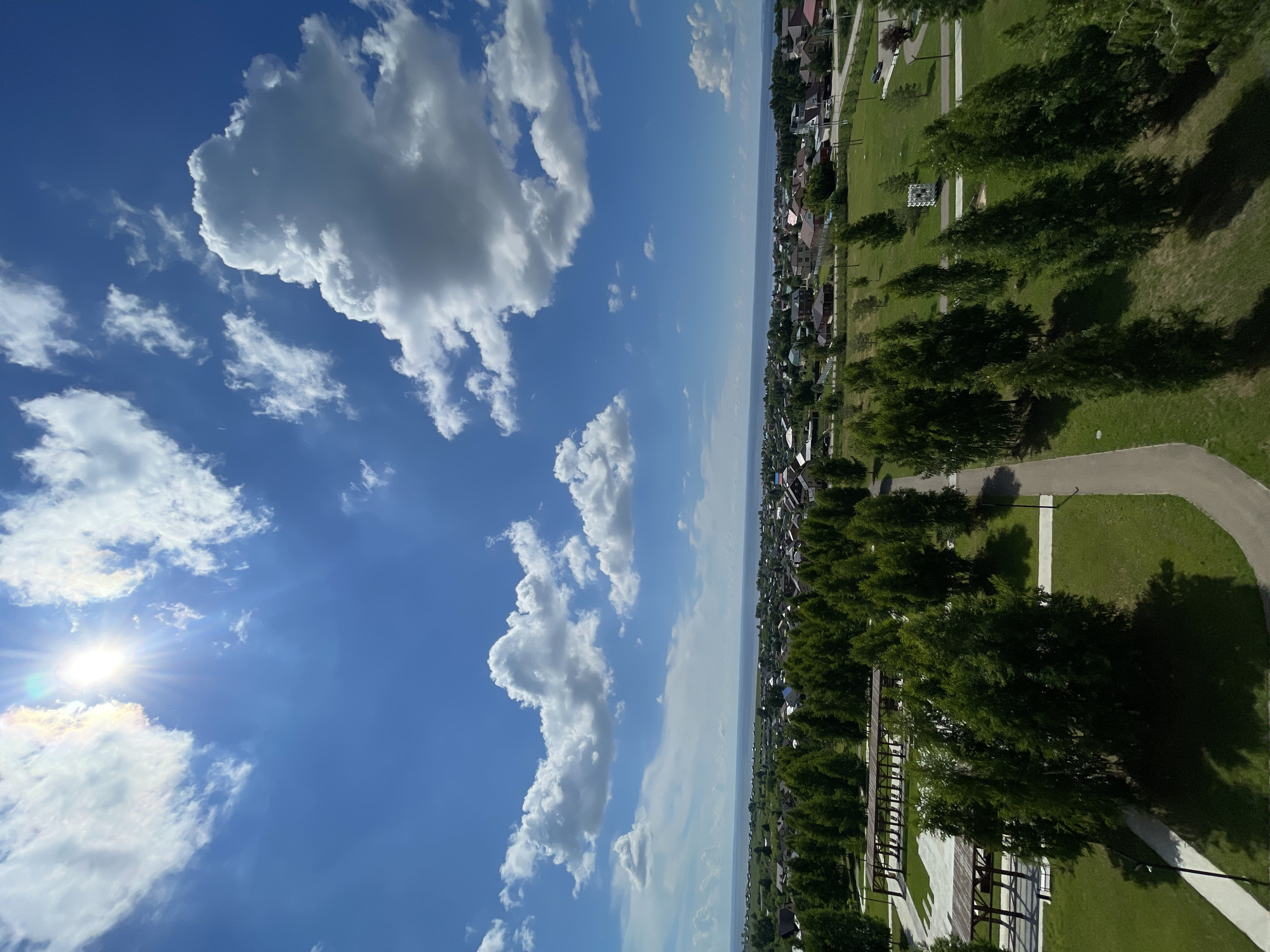 The image depicts a scenic view of the city of Laishevo, Russia, on a bright, sunny day. The foreground is dominated by a lush green park with manicured lawns and scattered trees. A paved pathway winds through the park, leading towards a residential area in the midground. The residential area consists of a collection of houses with red and brown roofs, interspersed with more trees. Beyond the houses, a wide body of water, likely the Kama River or a reservoir, stretches to the horizon. The background is a clear blue sky filled with fluffy white clouds. The sun is shining brightly, casting shadows on the ground and creating a halo effect around it. There are no people or animals visible in the scene.