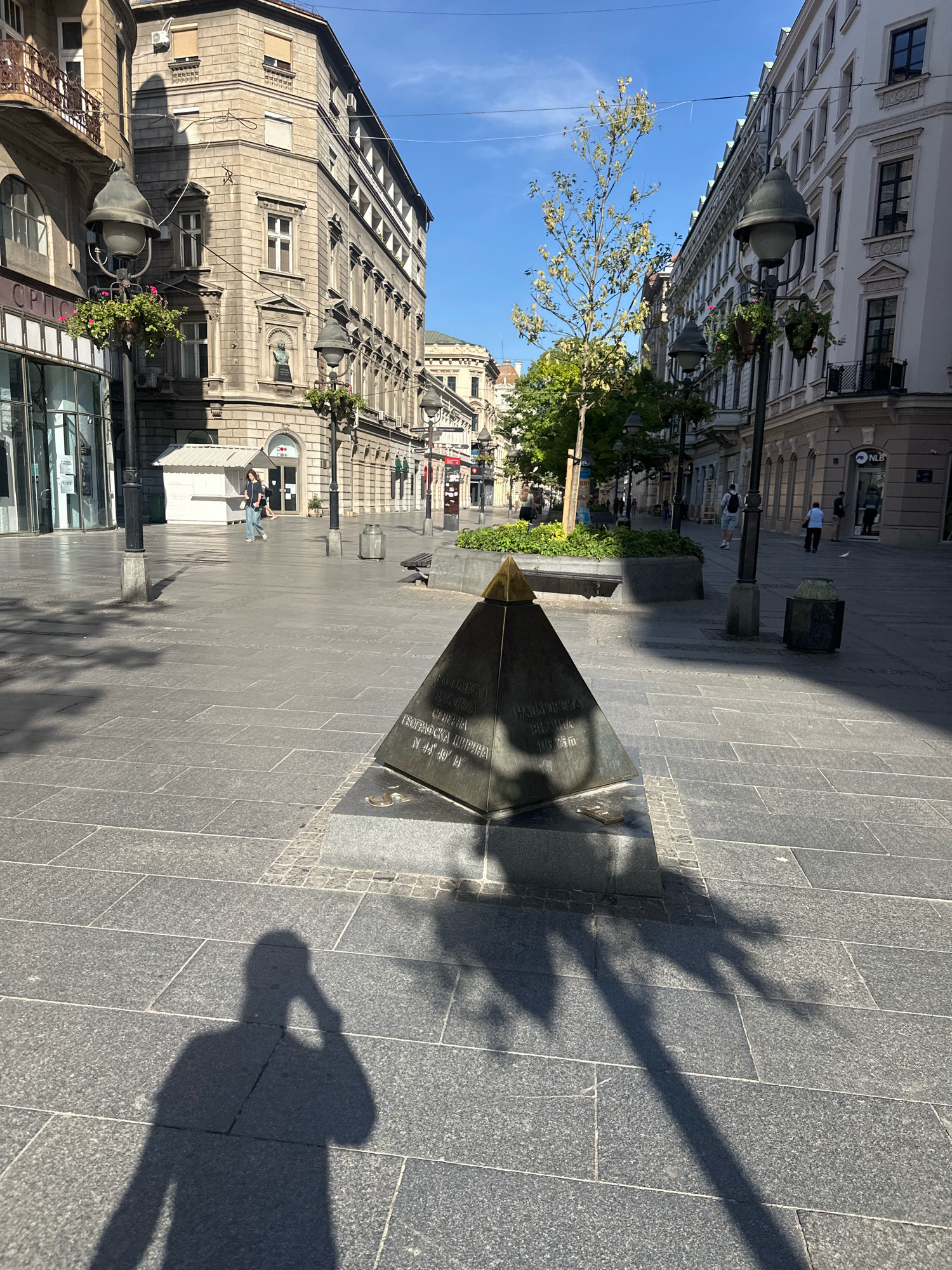 The image depicts a sun-drenched, pedestrianized street in Belgrade, Serbia. In the foreground, a pyramid-shaped monument with an inscription is visible, casting a long shadow. The inscription on the monument appears to be in Serbian and includes geographical coordinates and an elevation. The street is paved with large, dark grey stone tiles. The surrounding buildings are multi-story structures with classical architectural elements, typical of European city centers. Several streetlights with hanging flower baskets line the street. In the mid-ground, a few individuals are walking, and a lone pigeon is present. The sky is clear and blue, suggesting a pleasant day. The scene conveys a sense of quiet activity in an urban environment.