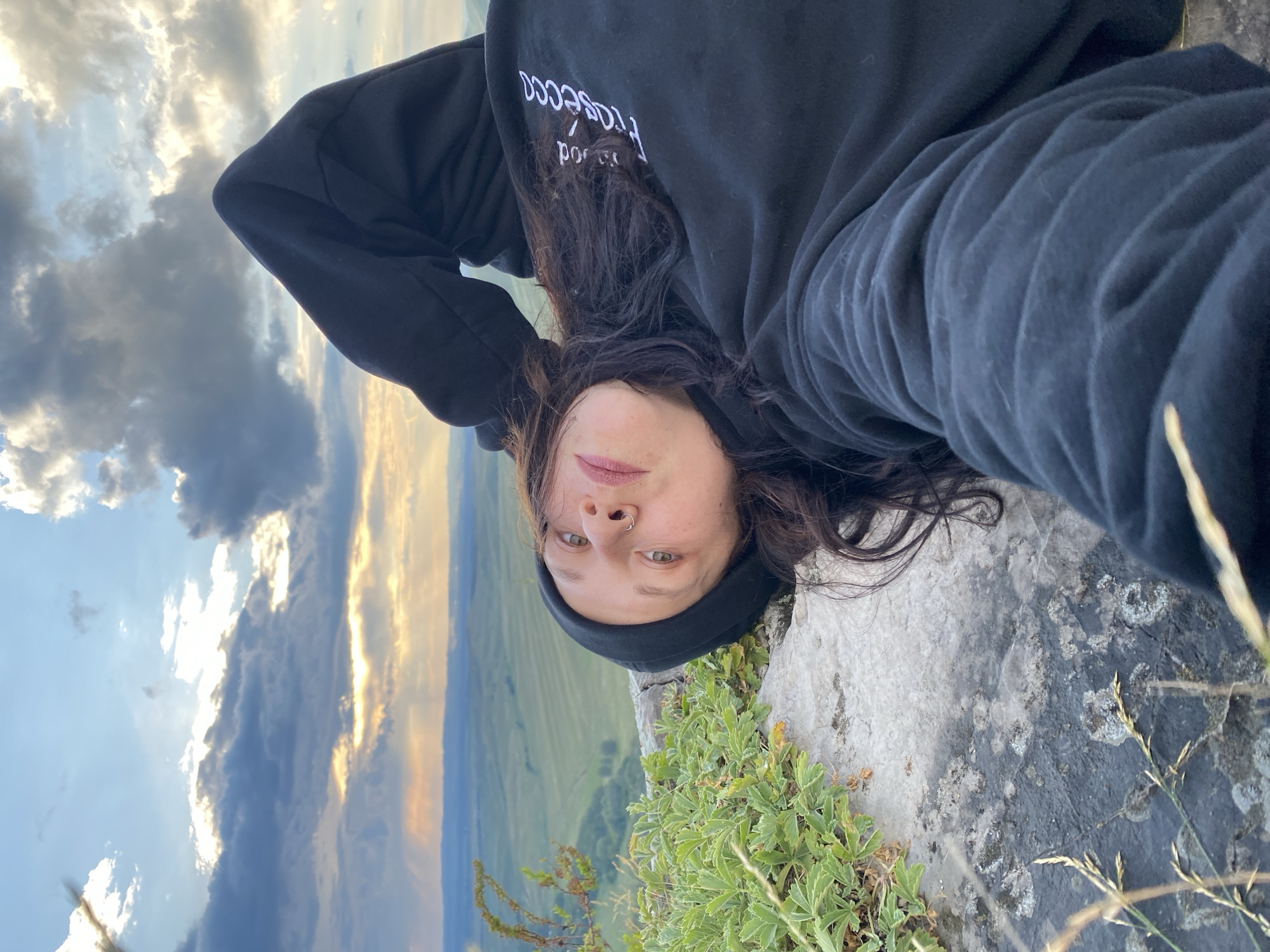A young woman with dark hair and a black beanie lies on a rock overlooking a vast, green landscape. She is wearing a black hoodie with white lettering on the sleeve. The sky above is filled with dramatic clouds, with rays of sunlight breaking through. The foreground shows the rocky terrain and some green plants. The location is in the village of Абзаново, Russia.
