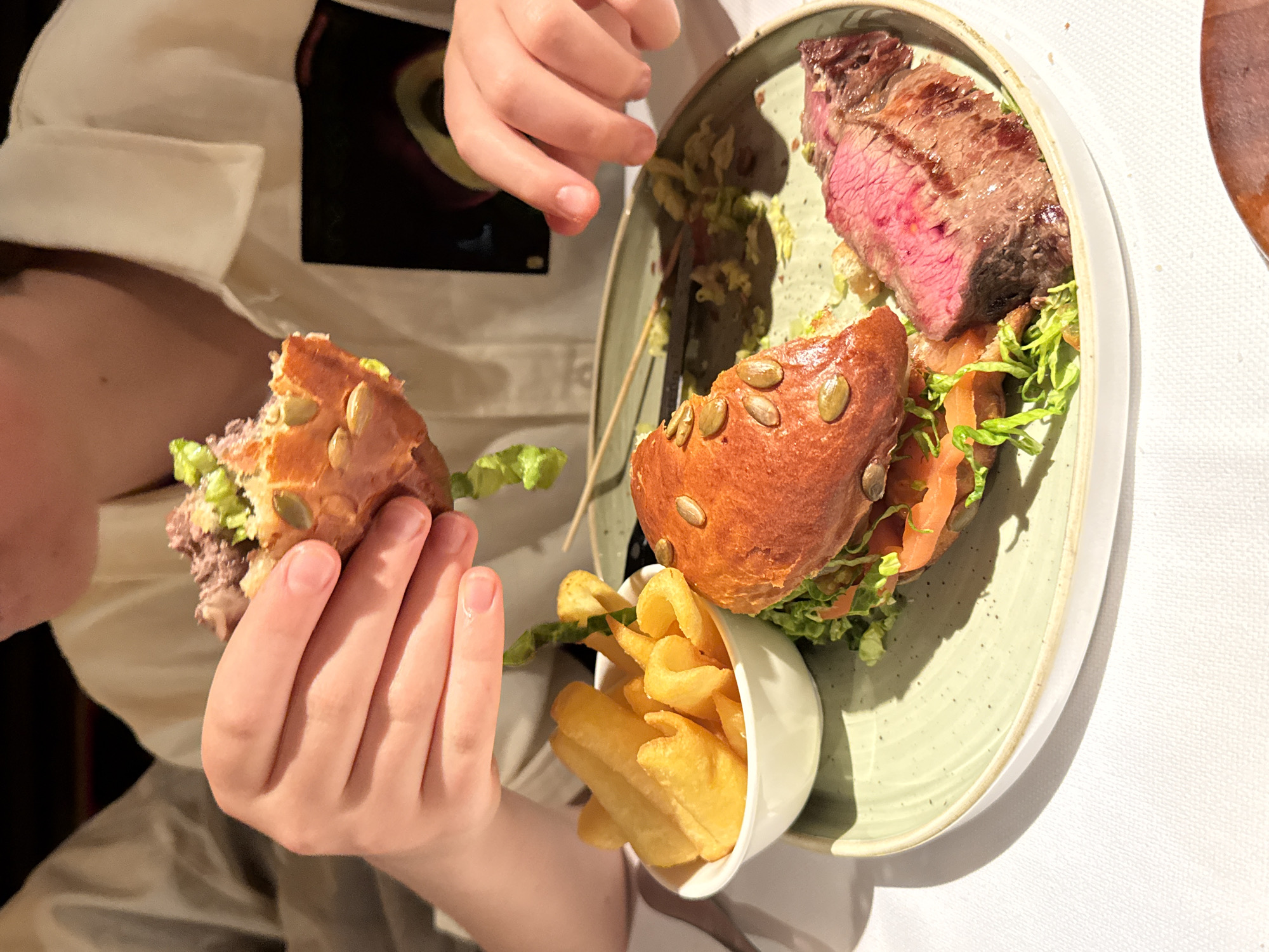 A child in a white shirt holds a partially eaten burger in their left hand. The burger has a seed-studded bun, meat, and lettuce. A plate contains the rest of the burger, a piece of medium-rare steak, lettuce, and a small white bowl filled with French fries. The plate is on a white tablecloth. The scene appears to be at a restaurant. There is no direct evidence of this image being from Milan, Italy.