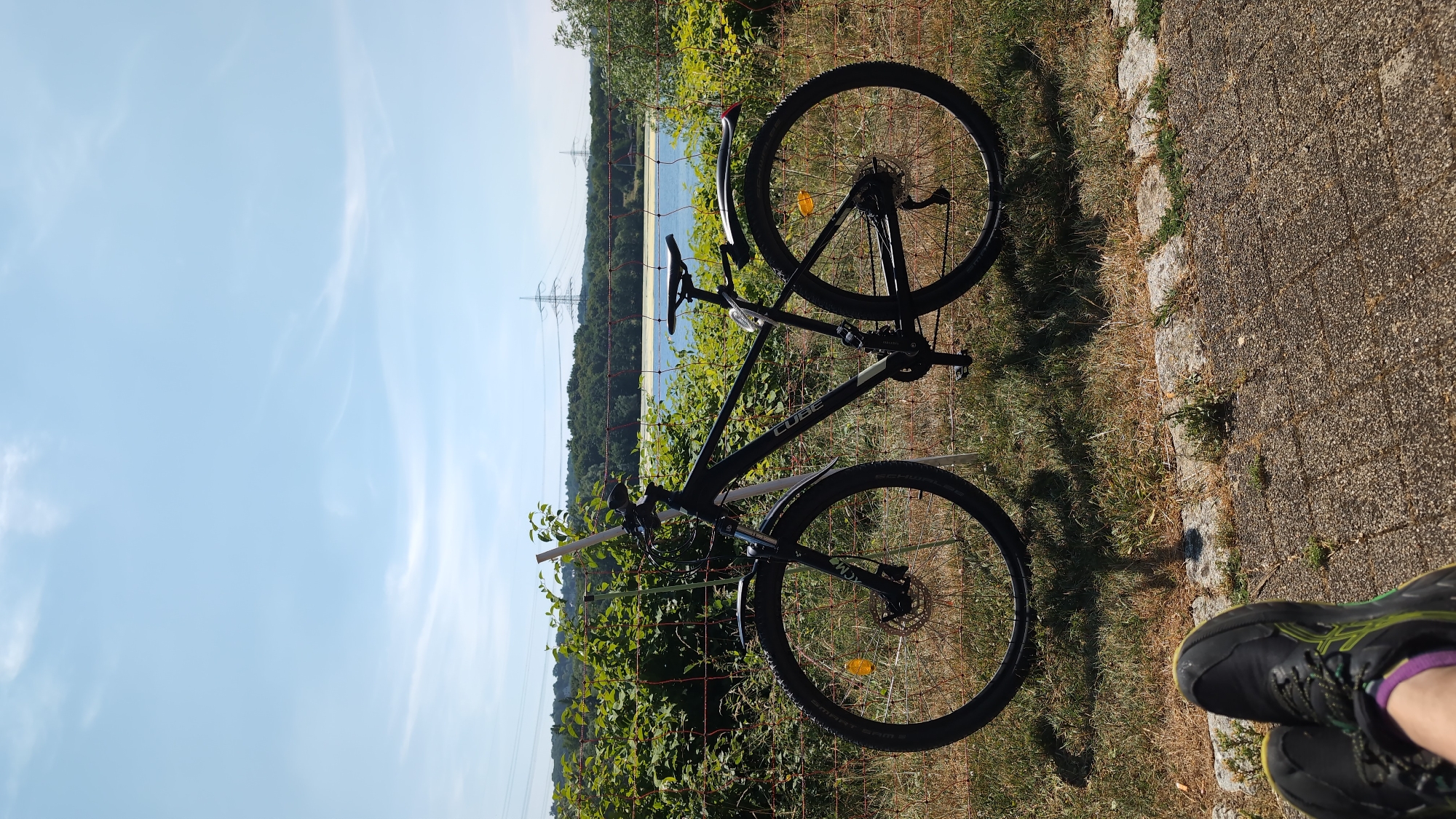A black Cube brand bicycle is parked upright on dry grass next to a red mesh fence. The bicycle has disc brakes, fenders on both wheels, and a kickstand. Beyond the fence is a body of water, likely a lake, surrounded by dense green foliage and trees. In the background, tall electrical transmission towers are visible against a clear blue sky with a few wispy clouds. In the foreground, a person's legs and feet, wearing dark athletic shoes with green accents and purple socks, are resting on a paved surface. The scene appears to be outdoors on a sunny day, suggesting a recreational area. While the image itself does not explicitly show any landmarks that would pinpoint it to Bochum, Germany, such a setting with a lake and power lines could be found in the outskirts or recreational areas of the city, such as near the Kemnader See or the large industrial areas with characteristic power line infrastructure.